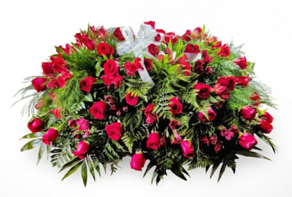 Red Rose Casket Spray  - Vivid red roses take center stage in this formal casket spray, a traditional sympathy arrangement designed to rest across a wooden casket and express deep love and respect at a funeral or memorial. The composition's broad, low silhouette and rich, solemn palette of red, green, and a white bow creates a dignified visual statement that helps the person you're honoring feel truly remembered.  Dense mixed greenery frames clusters of red roses and red alstroemeria, with tree fern tucked throughout for depth and texture; a white ribbon bow at the center provides a quiet contrast and a note of purity. Built to sit gracefully on a casket, this large, full spray conveys a timeless, comforting presence for mourners while honoring a life well lived.