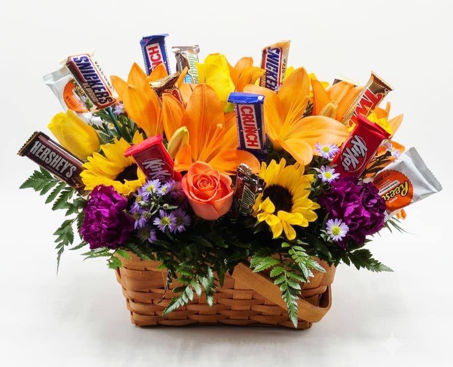Sweet and Vibrant Candy Flower Basket - Bright orange lilies and sunny yellow sunflowers make an immediate, joyful impression in this candy flower basket-perfect as a birthday arrangement, congratulations gift, or a casual thank-you surprise for someone who loves cheerful color. The low woven basket reads playful and approachable, so the recipient will feel genuinely celebrated when they see the vibrant mix and the little treats tucked inside.  This arrangement includes orange lilies, yellow sunflowers, orange roses, purple carnations, and lavender aster-style blooms arranged in a 12" tall low woven basket. Snack-size candy bars are nestled through the flowers for a gift-basket feel that combines floral beauty with a sweet surprise. The warm oranges and yellows are balanced by deep purple accents for lively contrast and visual texture.  Send this to the person you want to brighten instantly-it's made to convey congratulations, birthday wishes, or simple thanks. 