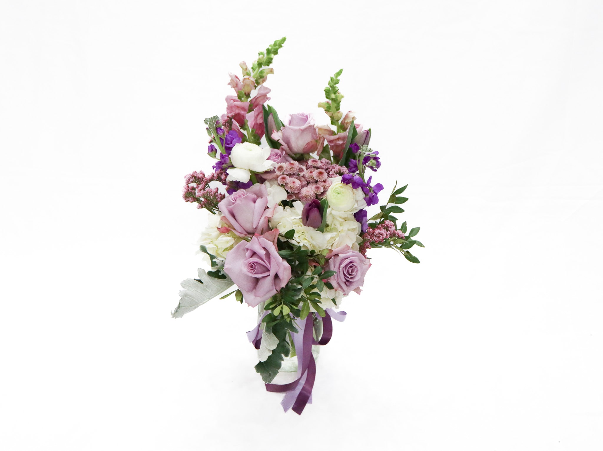 Verona - Lavender Whisper is an elegant front-facing floral arrangement designed in a clear glass vase featuring a sophisticated blend of purple and white blooms. This garden-inspired bouquet includes roses, tulips, hydrangeas, snapdragons, delphinium, and seasonal greenery arranged with soft texture and graceful height. The fresh mix of lavender, ivory, and soft green tones creates a timeless and romantic design perfect for birthdays, sympathy gestures, anniversaries, thank you gifts, or adding beauty to any space.