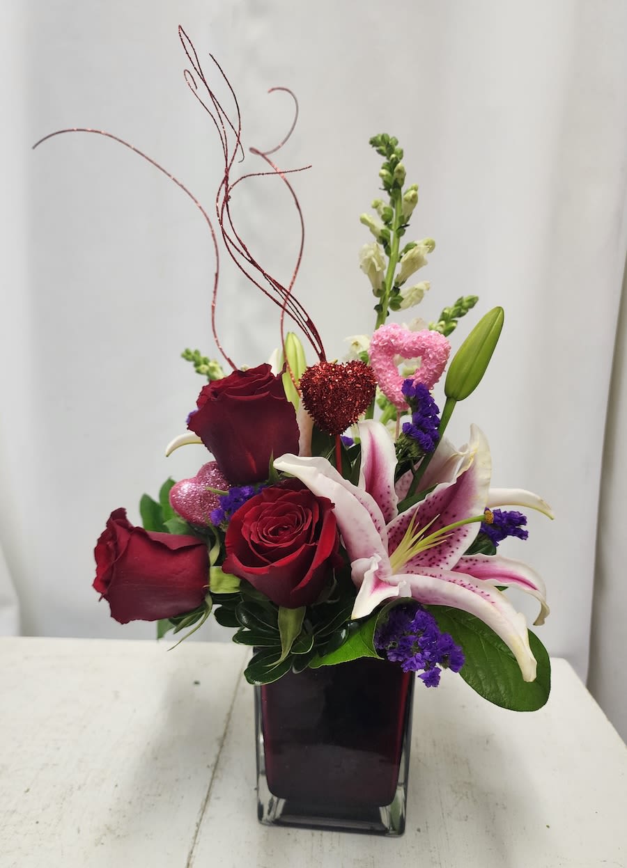 Red roses and pink lilies in a square glass vase with heart picks