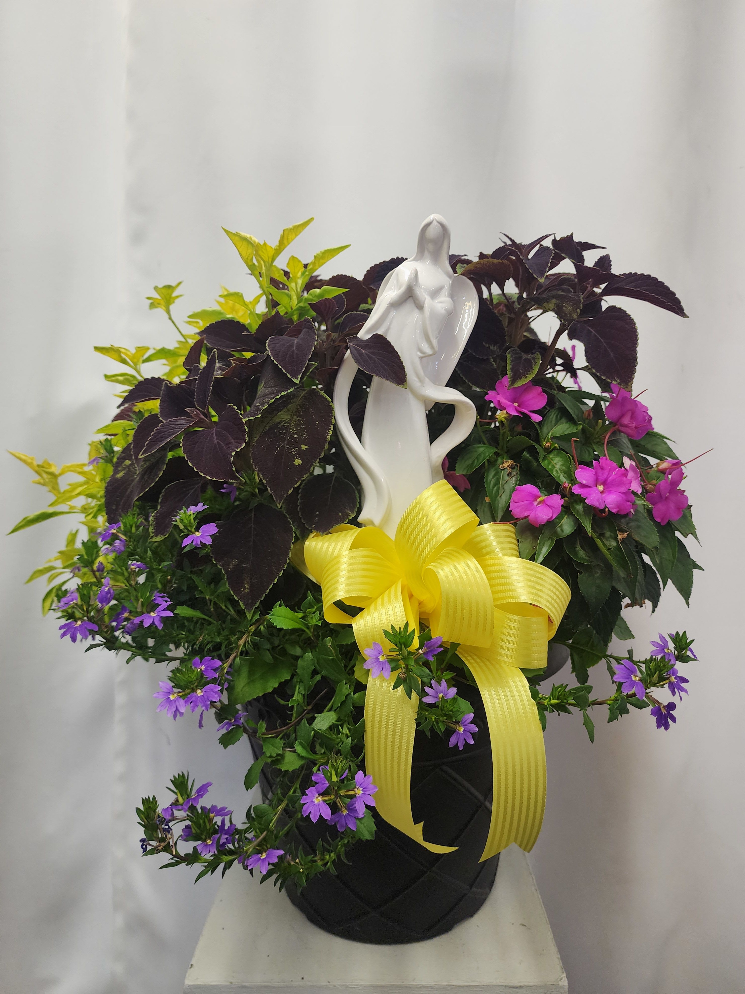 Potted mixed flowering plant arrangement with a white figurine and yellow ribbon