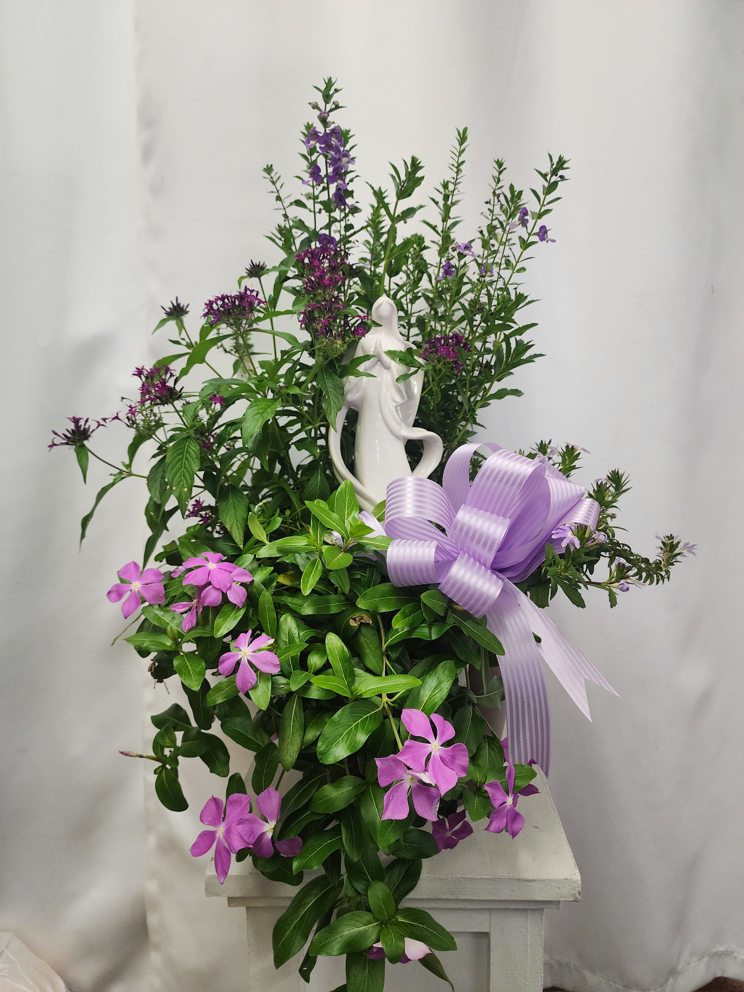 Purple flowers and greenery arranged around a white figurine with a lavender ribbon bow.