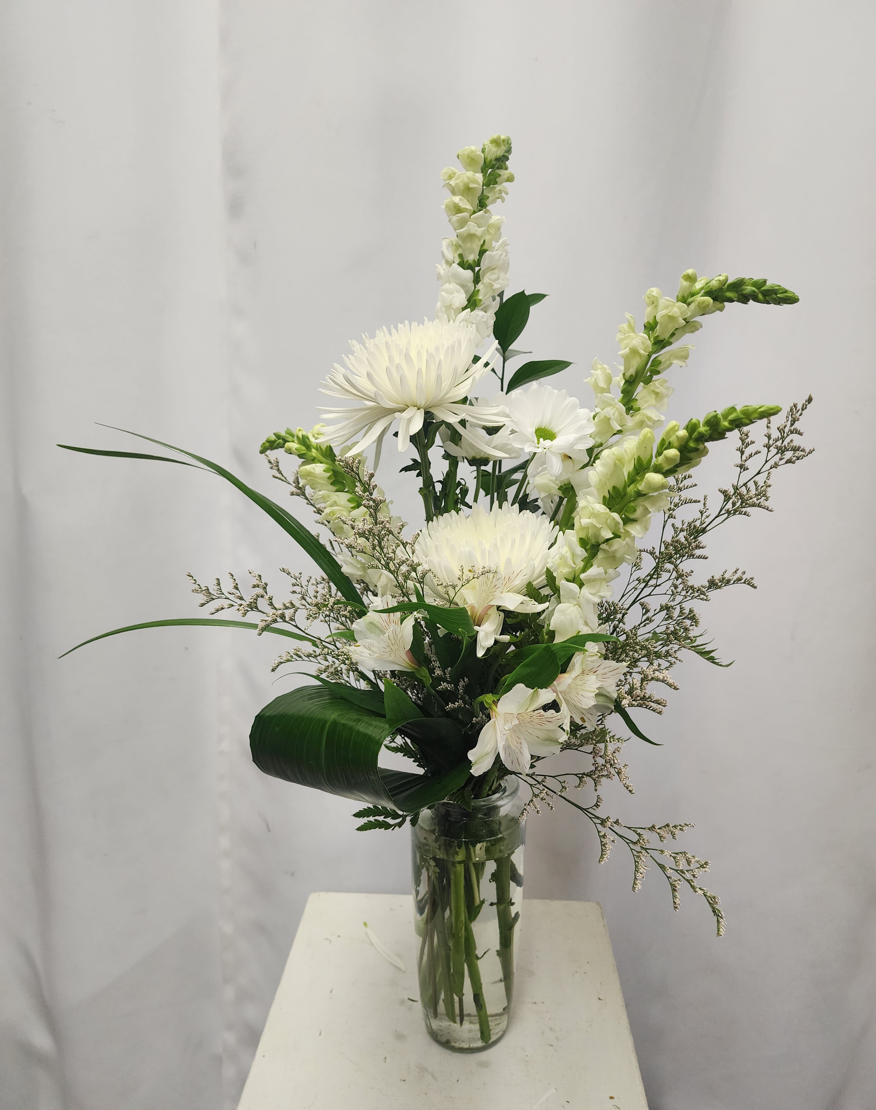 Whimsical in White - “Whimsical in White” captivates with a classic palette and modern flair. This tall, graceful piece features snapdragons, spider mums, alstroemeria, and cheerful daisies, all artfully arranged with creative greenery in a striking clear glass vase. The result is a luminous symphony of texture and light—perfect for celebrating new beginnings, expressing heartfelt sympathies, or simply brightening any space with timeless elegance. 