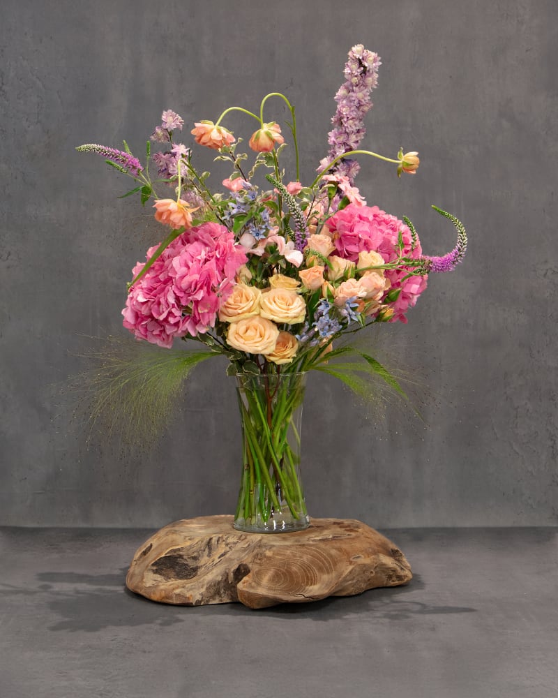 Tall bouquet of pink hydrangeas, peach roses, and lavender flowers in a glass vase
