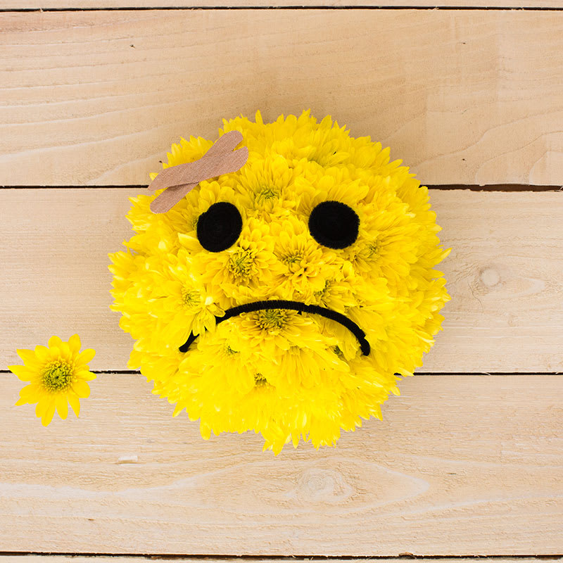 Round yellow chrysanthemum arrangement shaped like a sad face with bandage on forehead on wooden surface