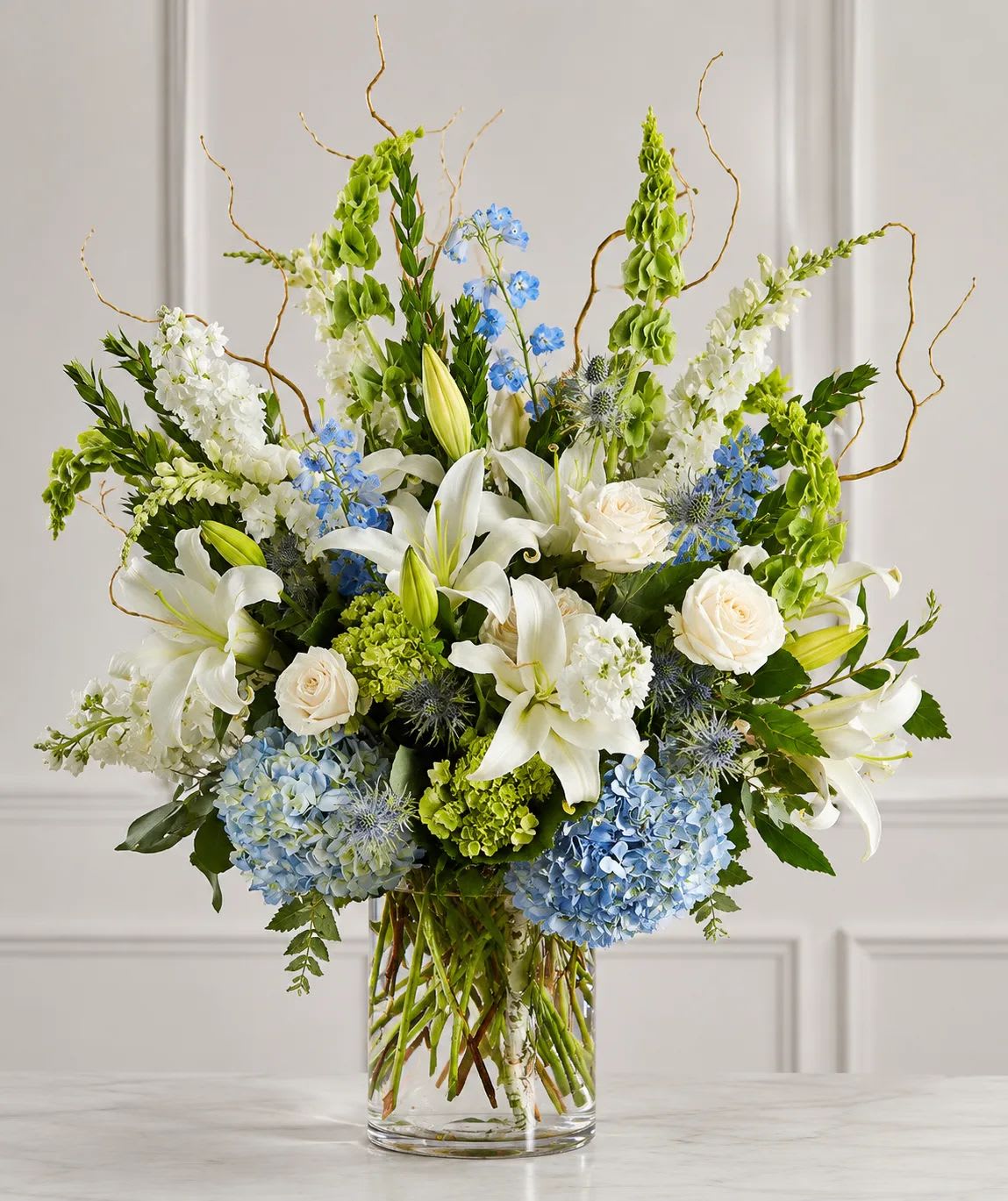 White lilies, roses, blue flowers, and green blooms in a clear glass vase