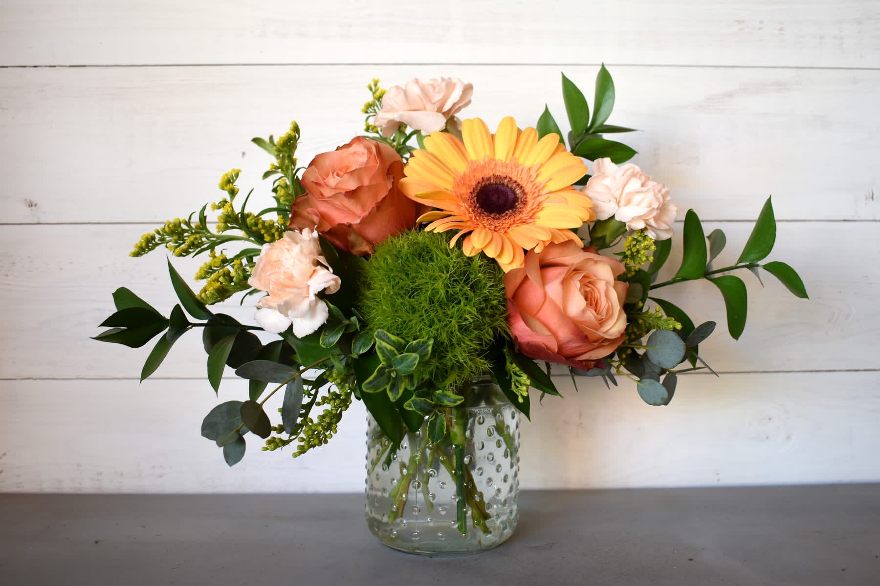 Peach roses, pale carnations, a yellow gerbera daisy, and a green pompom bloom in a clear glass jar.