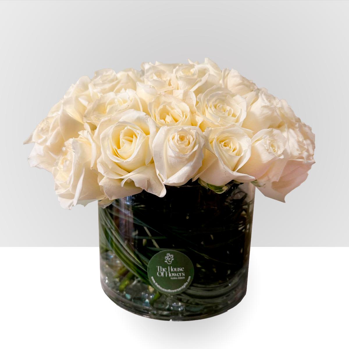  Ivory Garden Luxe - Three Dozen Roses by The House of Flowers - A luxurious arrangement featuring three dozen premium ivory roses, beautifully designed in a clear glass vase. The stems are elegantly displayed through the glass and accented with decorative stones, creating a refined and modern presentation.  Fresh, assorted foliage adds natural texture and depth, enhancing the soft beauty of the roses and giving the arrangement a full, lush look.  This elegant design makes a stunning statement and is perfect for special celebrations, sympathy gestures, anniversaries, or sophisticated home décor. Classic, graceful, and timeless, it brings warmth and elegance to any space.  Arrangement Details: Three dozen white roses in a glass cylinder base. 