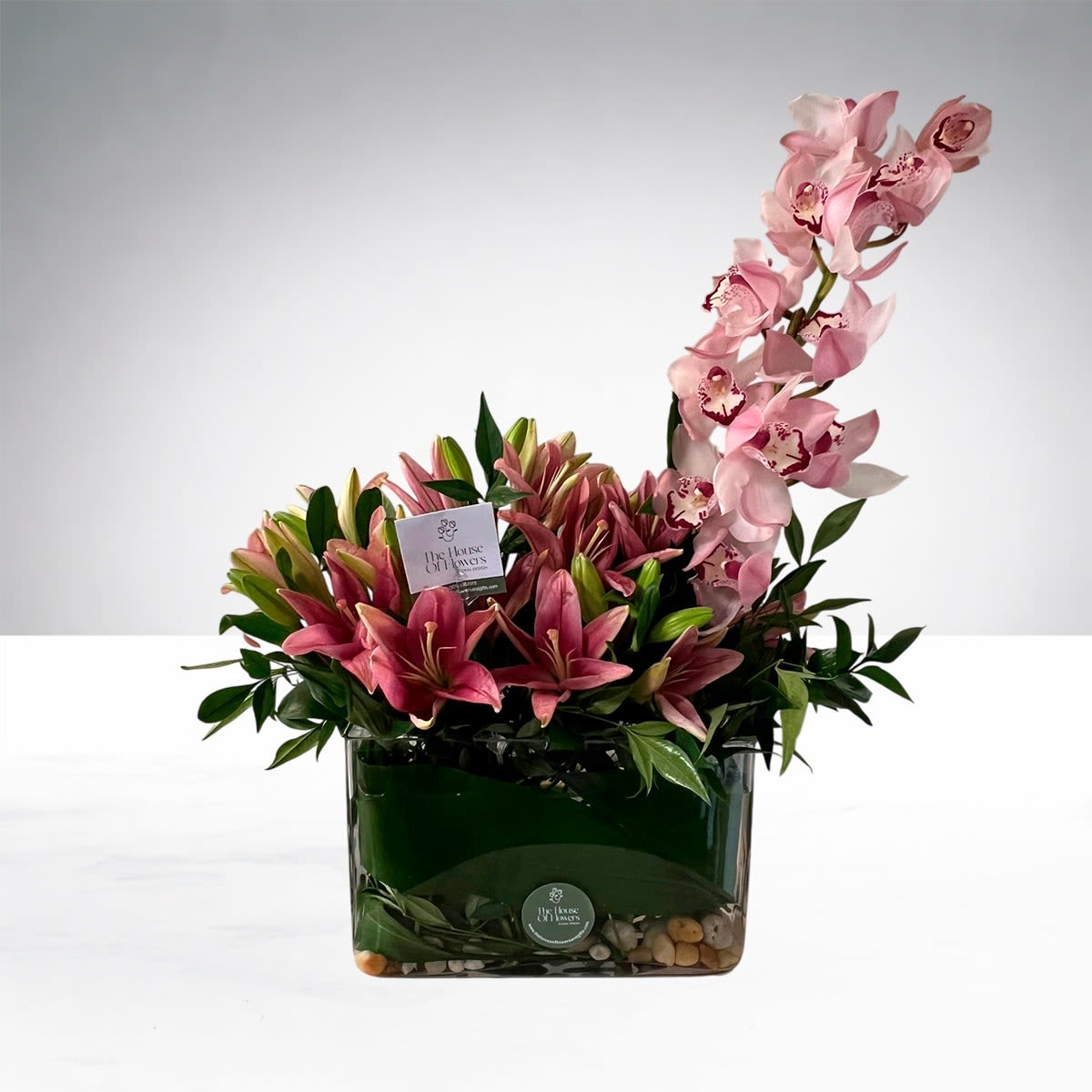 Blush Orchid Elegance by The House of Flowers - A sophisticated floral arrangement featuring premium pink lilies paired with a graceful orchid stem that adds an elegant and luxurious touch. Designed in a modern glass vase with natural decorative stones, fresh greenery, and a refined contemporary style, this arrangement is perfect for celebrating life’s most special moments.  Flower colors may vary slightly based on seasonal availability.
