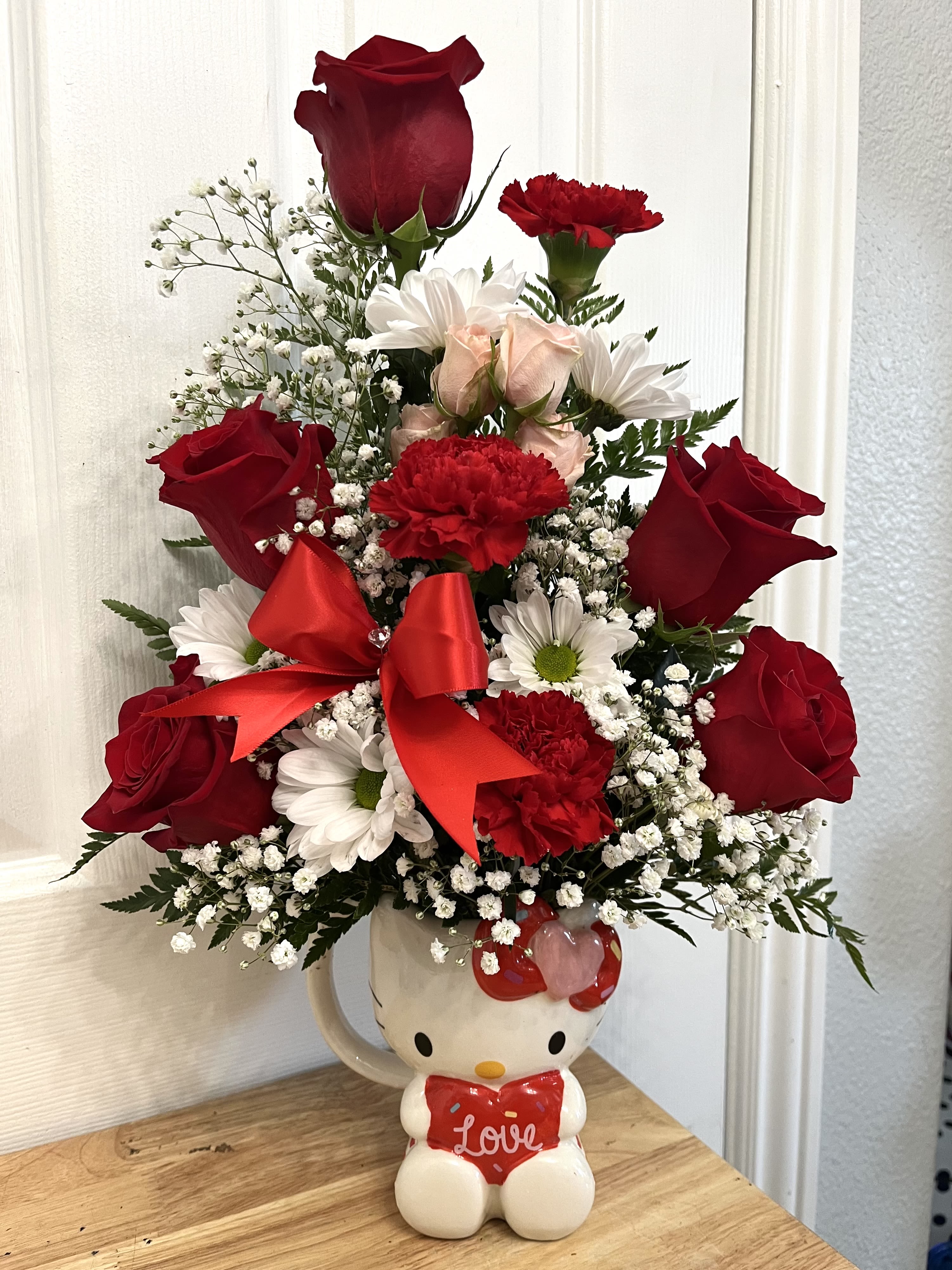 Red roses and white daisies arranged in a Hello Kitty mug with a red bow.