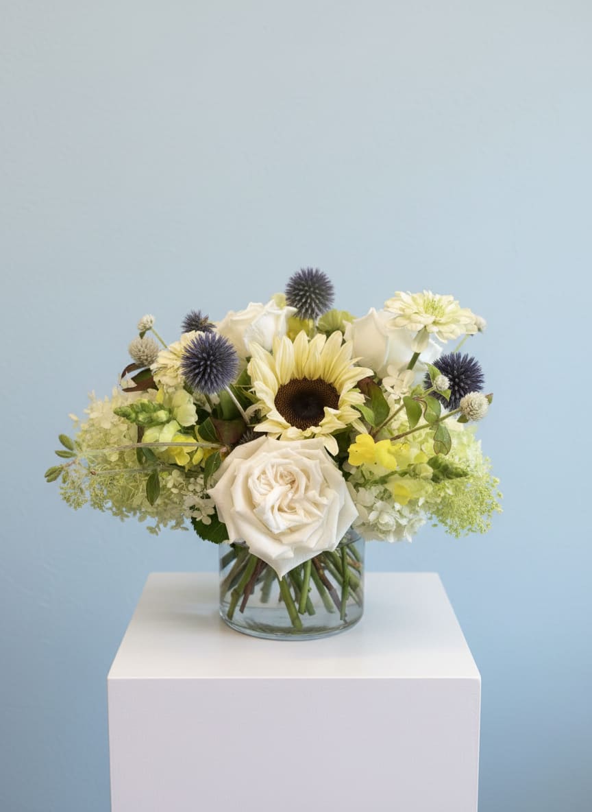 White roses and a sunflower in a glass vase with blue thistle accents
