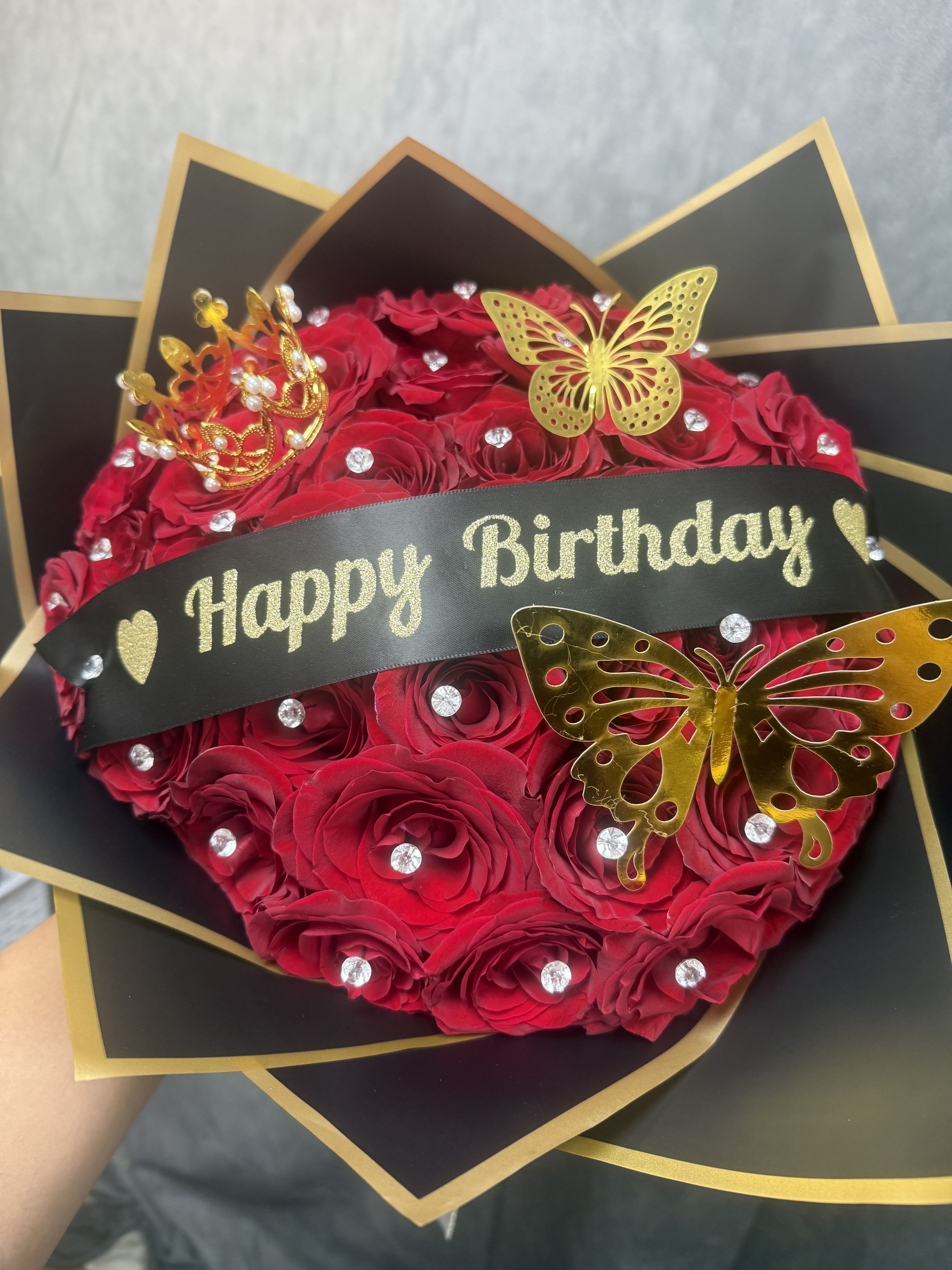 The Birthday Queen Bouquet - Fit for royalty, our Birthday Queen Bouquet features luxurious red roses adorned with sparkling crystal accents, elegant butterflies, and a charming crown centerpiece. Wrapped in sophisticated black and gold paper and finished with a festive birthday ribbon, this stunning bouquet is designed to make the birthday queen feel celebrated, cherished, and truly special on her big day. 