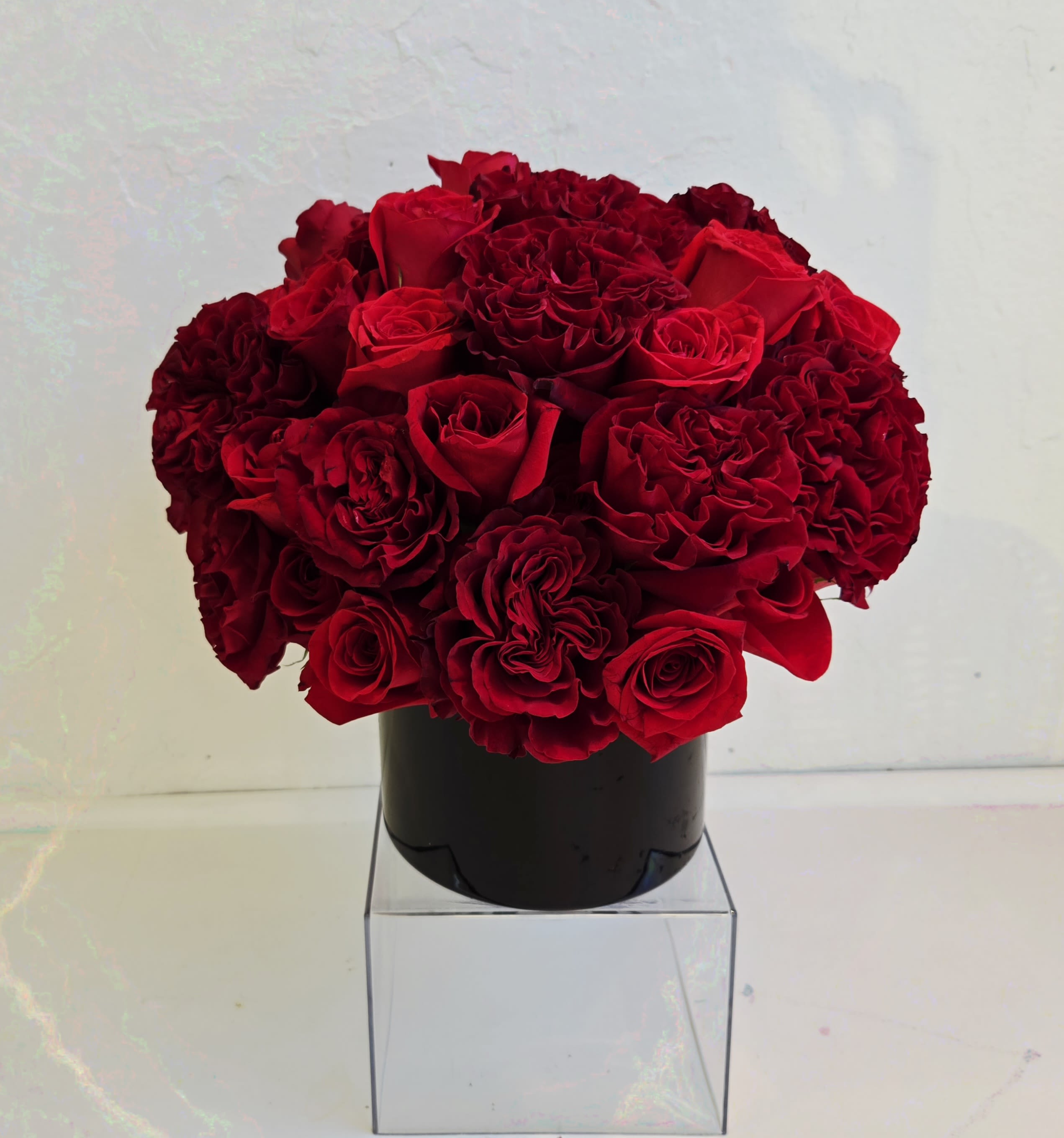 Compact arrangement of red roses and carnations in a black vase on a clear cube stand
