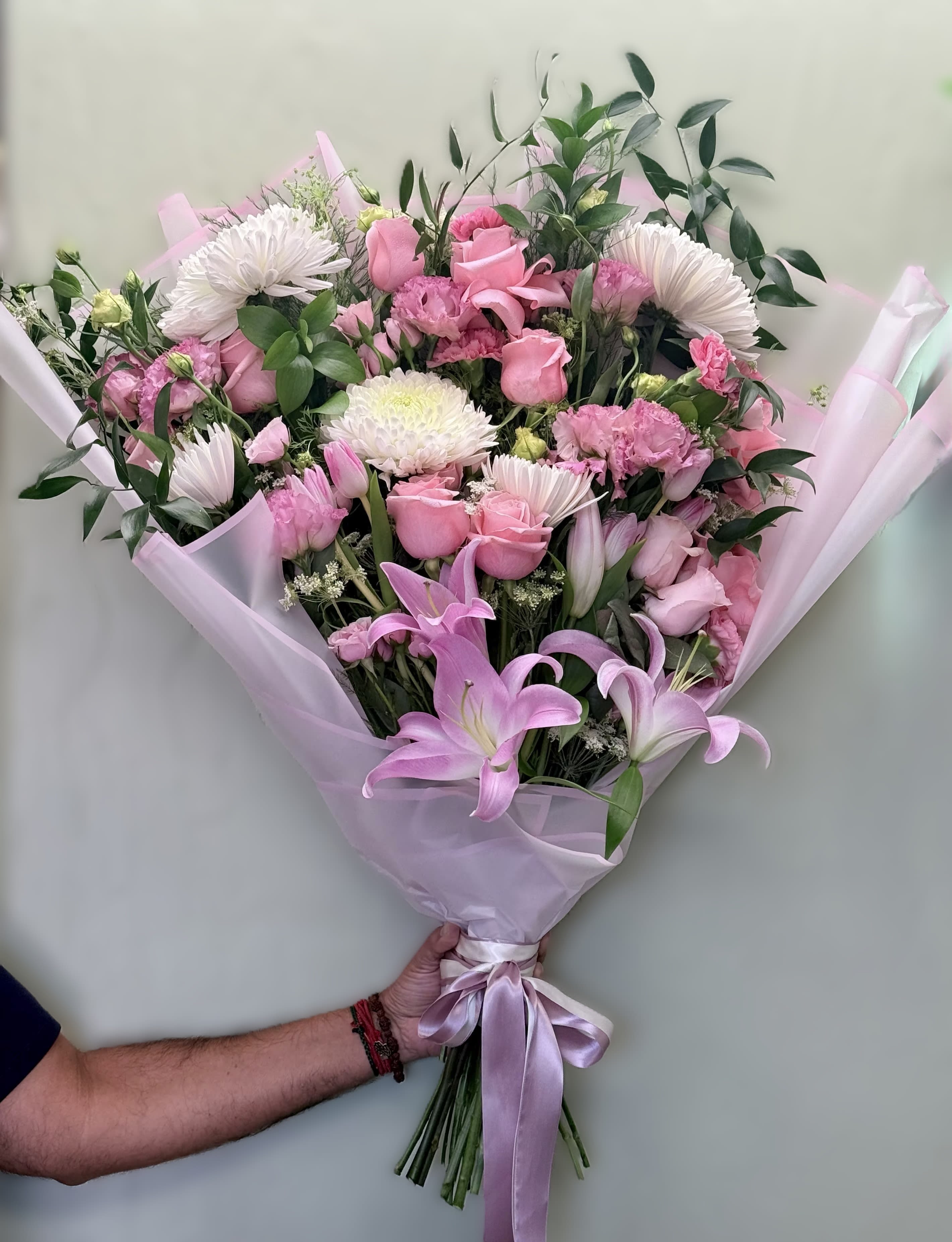 The Regal Romance by The Flower Spot™ - A lavish, hand-tied bouquet that reads like a love letter in soft pastels-perfect as a birthday bouquet, anniversary roses alternative, or a thoughtful "thinking of you" arrangement. The recipient will feel genuinely celebrated the moment they see the lush mix of pink roses and stately lilies framed by white daisy-like blooms, lavender filler, and fresh green accents-a romantic presentation that says affection and elegance.  Carefully curated by our designer, this large arrangement layers open blush roses, lily buds for height, chrysanthemums, lisianthus, alstroemeria, and airy filler florals to create a full, textured silhouette. Pale pink wrapping paper and a dual ribbon-satin with a gold-edged trim-give it a polished, gift-ready finish. The bouquet arrives densely packed and wide-spread for an abundant look; expect a generous, hand-tied size that makes a striking table centerpiece or focal gift.  Ideal for anniversaries, Mother's Day, birthdays, or simply to make someone feel truly seen, this arrangement is arranged to hold water well until it's transferred to a vase. For best longevity, recut stems, change water daily, and keep away from direct heat-ordering is available for same-week local delivery and careful gift wrapping ensures it arrives ready to impress.