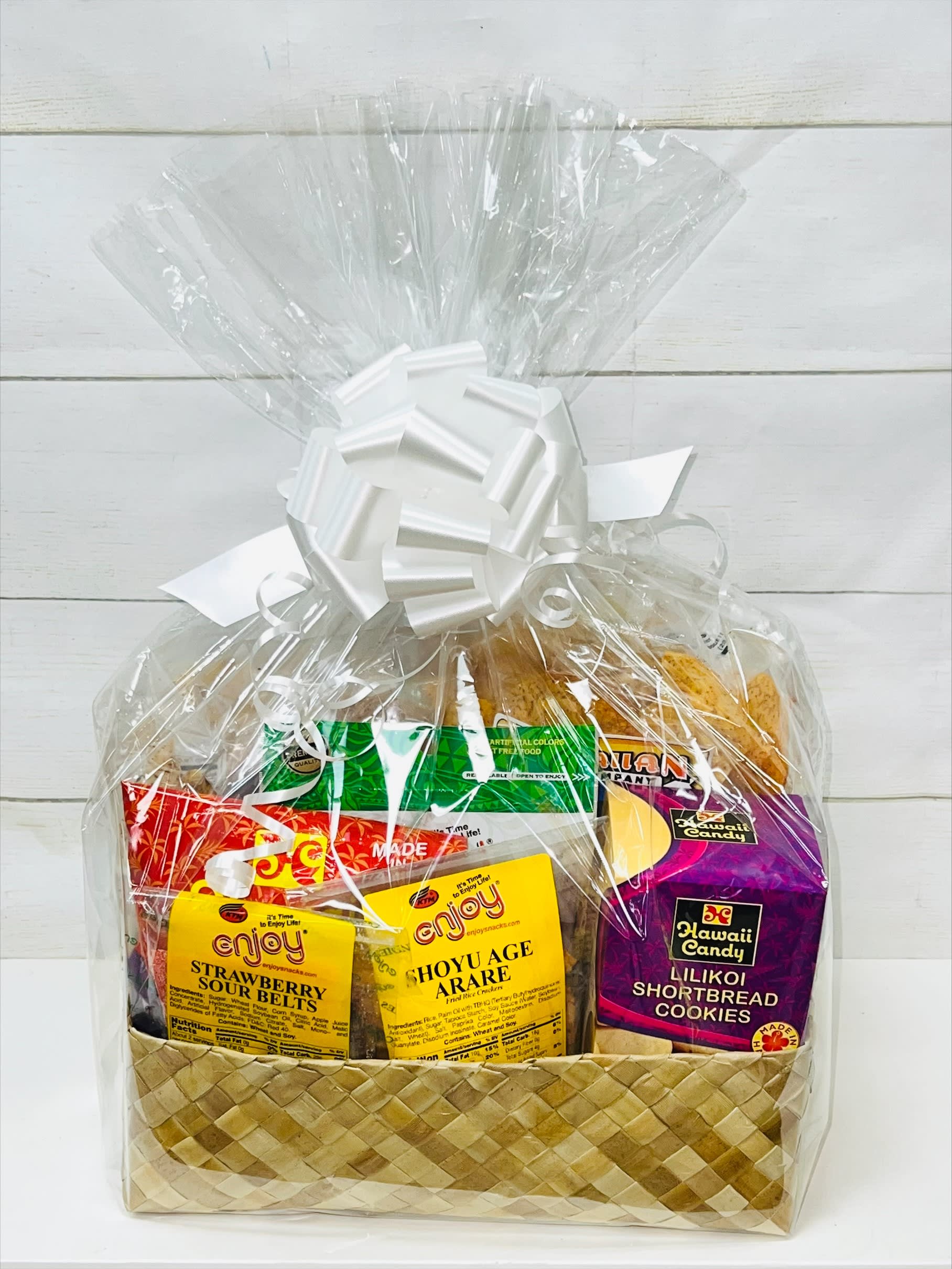 Woven gift basket wrapped in clear cellophane, filled with assorted colorful snack packages and topped with a white bow.