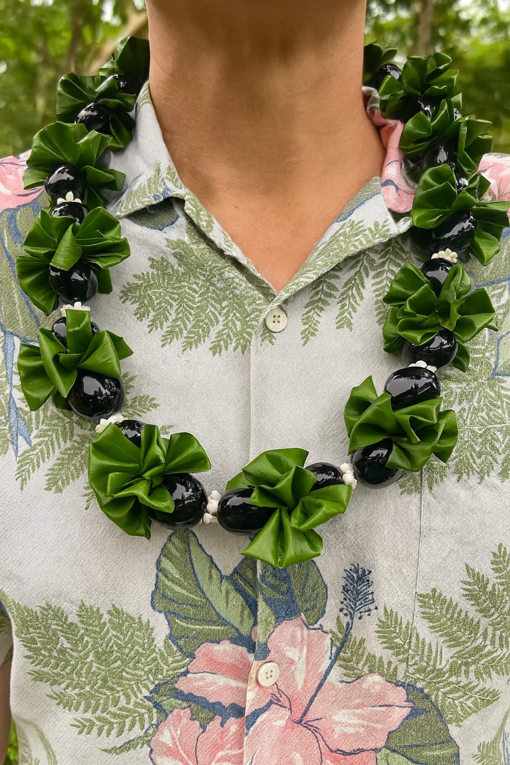 Glossy black kukui nut lei with green ribbon clusters worn over a tropical shirt.