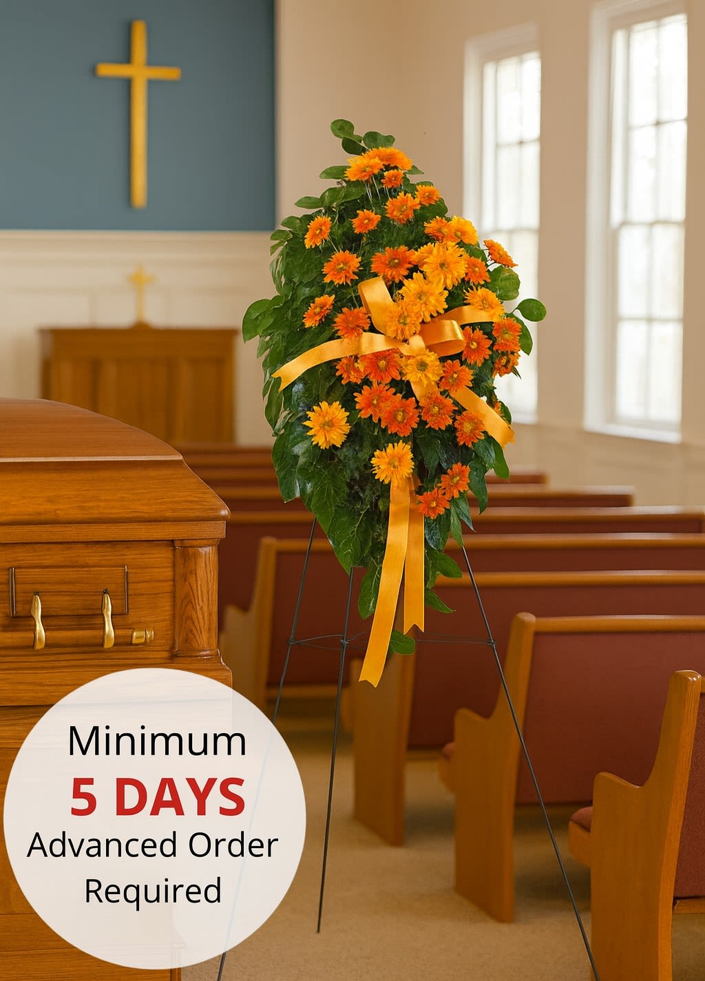 Standing spray of bright orange daisies with yellow bow on an easel beside a wood casket in a chapel.