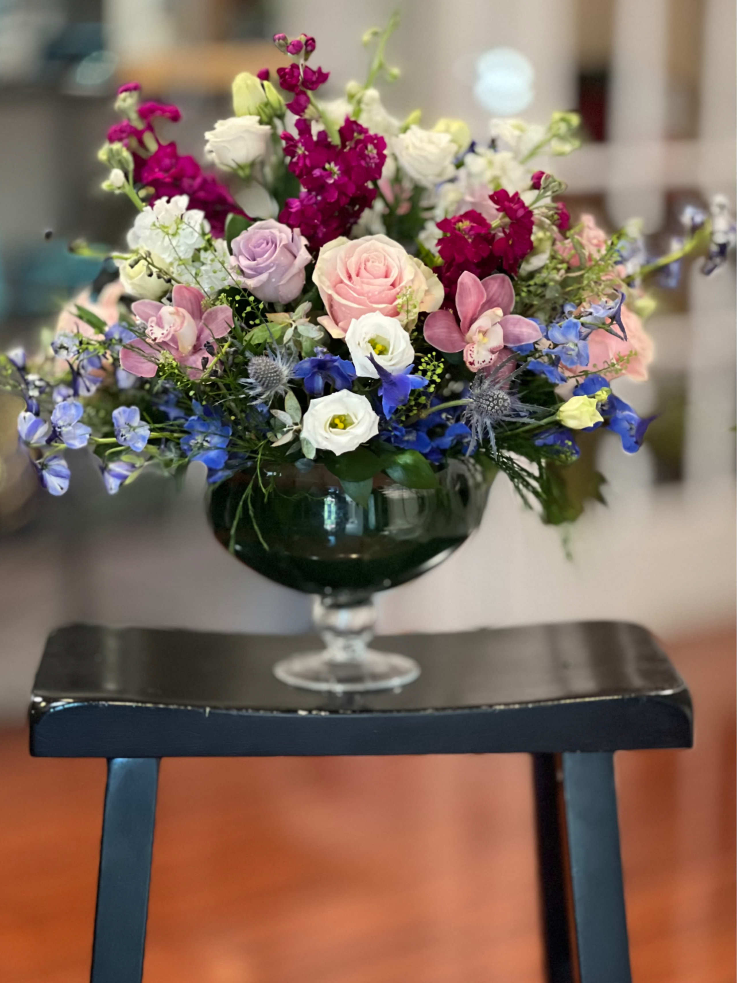 Jewel Tones - A lush, rounded mixed bouquet in jewel tones-soft pink and white roses clustered at the center with lavender lisianthus, sky-blue delphinium, and vivid magenta spikes-presented in a dark green glass goblet vase. This flower arrangement for birthdays, anniversaries, or a cheerful thank-you or just-because gift reads romantic and celebratory, the layered color pops creating an immediate "made for them" moment that will make the recipient feel truly seen.  Perfect for someone who loves bright, elegant flowers-send it for a birthday, anniversary, or to brighten an ordinary day. Vase-arranged for immediate enjoyment; refresh water and recut stems every few days to keep blooms looking their best.