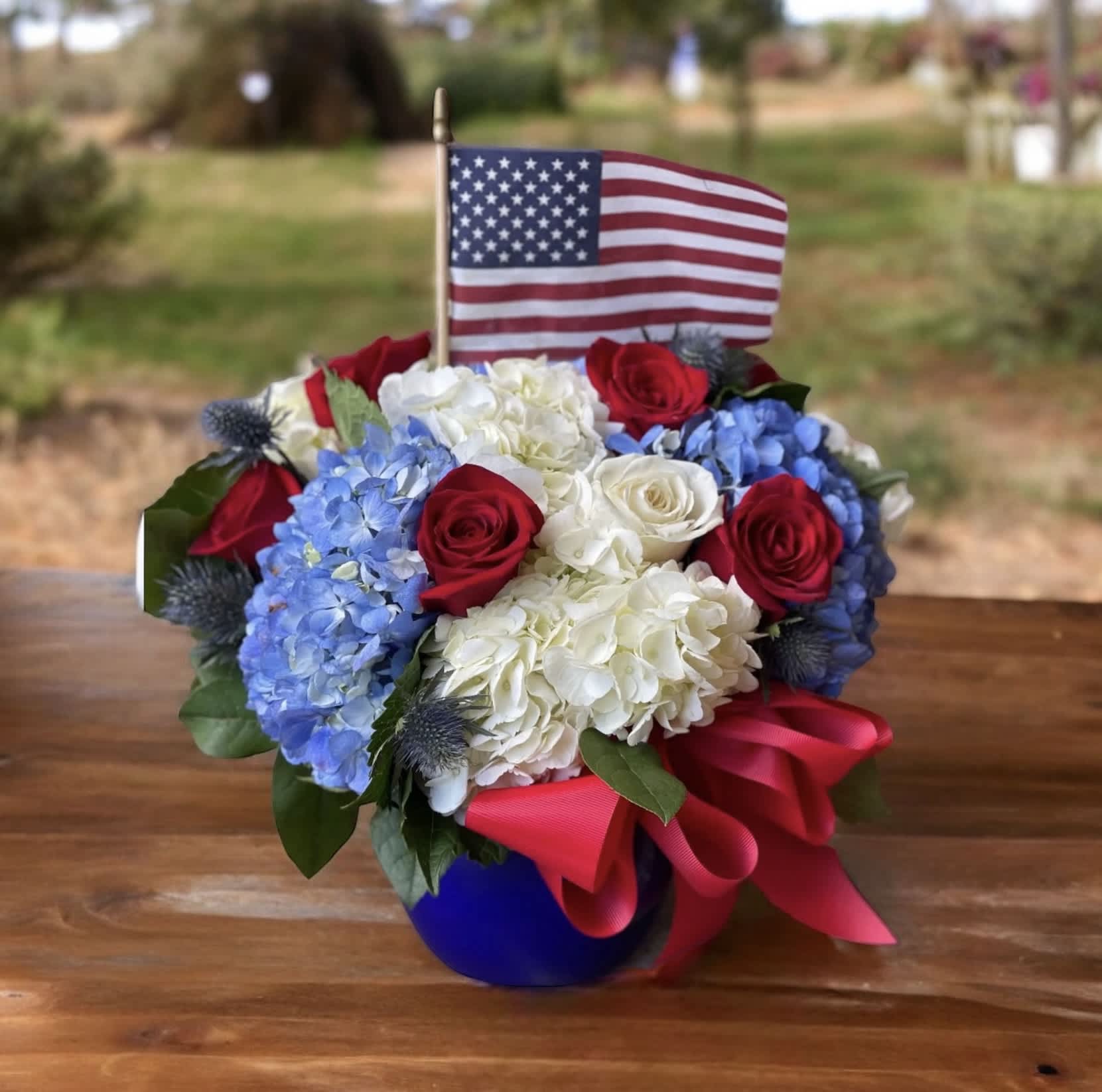 Americana - Americana – Patriotic Red, White &amp; Blue Flower Arrangement | Wilmington, DE Local Florist  Celebrate with pride and elegance with The Americana, a stunning patriotic floral arrangement designed by Petals Flowers and Fine Gifts, your trusted local florist in Wilmington, Delaware. This striking design features lush white hydrangeas, vibrant blue hydrangeas, classic red and white roses, and bold blue thistle, all beautifully arranged in a charming blue pitcher. A red satin ribbon and an American flag add the perfect finishing touches, making this arrangement ideal for Memorial Day, Independence Day, Veterans Day, or any occasion that calls for honoring our nation.  We offer same-day flower delivery in Wilmington, DE, and to nearby areas including Hockessin, Newark, Glen Mills, Chadds Ford, Christiana, and Kennett Square.  Why Choose Us:  Locally owned boutique florist dedicated to the Wilmington community Custom-designed, premium flower arrangements with exceptional attention to detail Satisfaction guaranteed with every order Enjoy tax-free flower delivery on all deliveries Order The Americana today from Petals Flowers and Fine Gifts and send a meaningful, patriotic floral tribute that truly stands out.