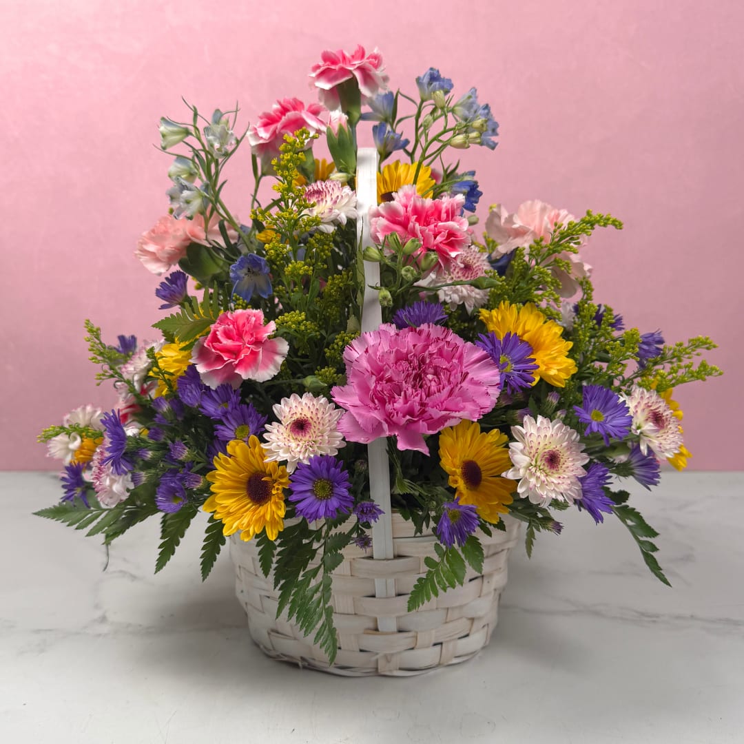 Celebrating You Basket - Our Celebrating You Basket is a joyful floral arrangement, a great choice for a birthday bouquet or spring gift that also reads perfectly as a Mother's Day, thank-you, or "just because" surprise. The garden-style mix of textures and mixed-color blooms creates an upbeat, celebratory impression that will make the person you're thinking of feel truly seen and appreciated.  Carefully arranged for a dense, rounded profile with a white handle, the Celebrating You Basket pairs colorful carnations, blue delphinium, and lavender chrysanthemums and button mums with small filler blooms to add depth and movement. The palette gives the design both softness and pop; the taller blue delphinium lifts the composition for a lively, airy silhouette. Ideal for spring gifting, birthdays, or a thoughtful thank-you, it arrives ready to display in its woven basket, simply add water every couple of days to keep blooms at their best.