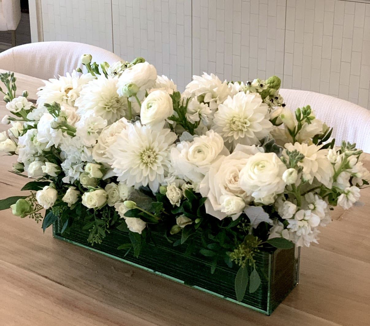 Low rectangular arrangement of white roses, dahlias, and other blooms in a green glass vase