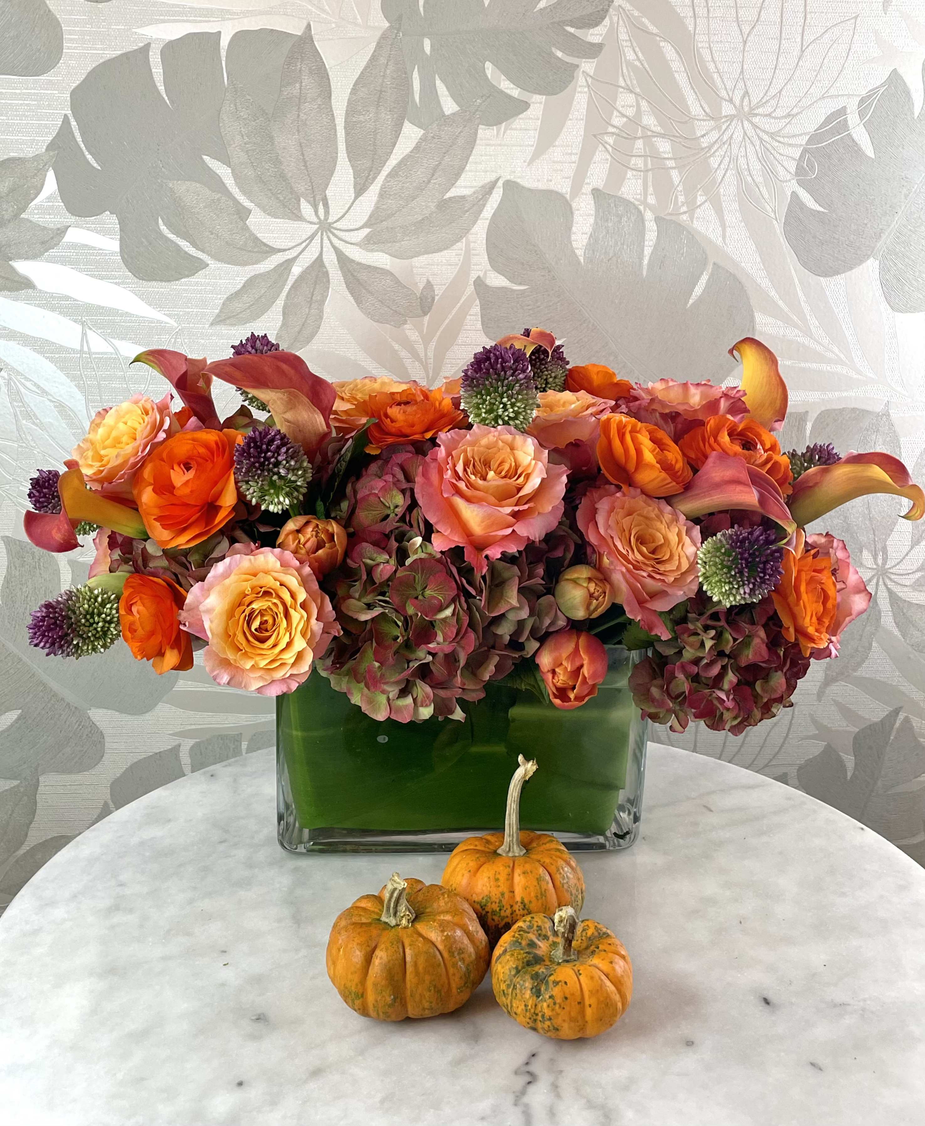 Orange and pink floral arrangement in a square glass vase with small pumpkins