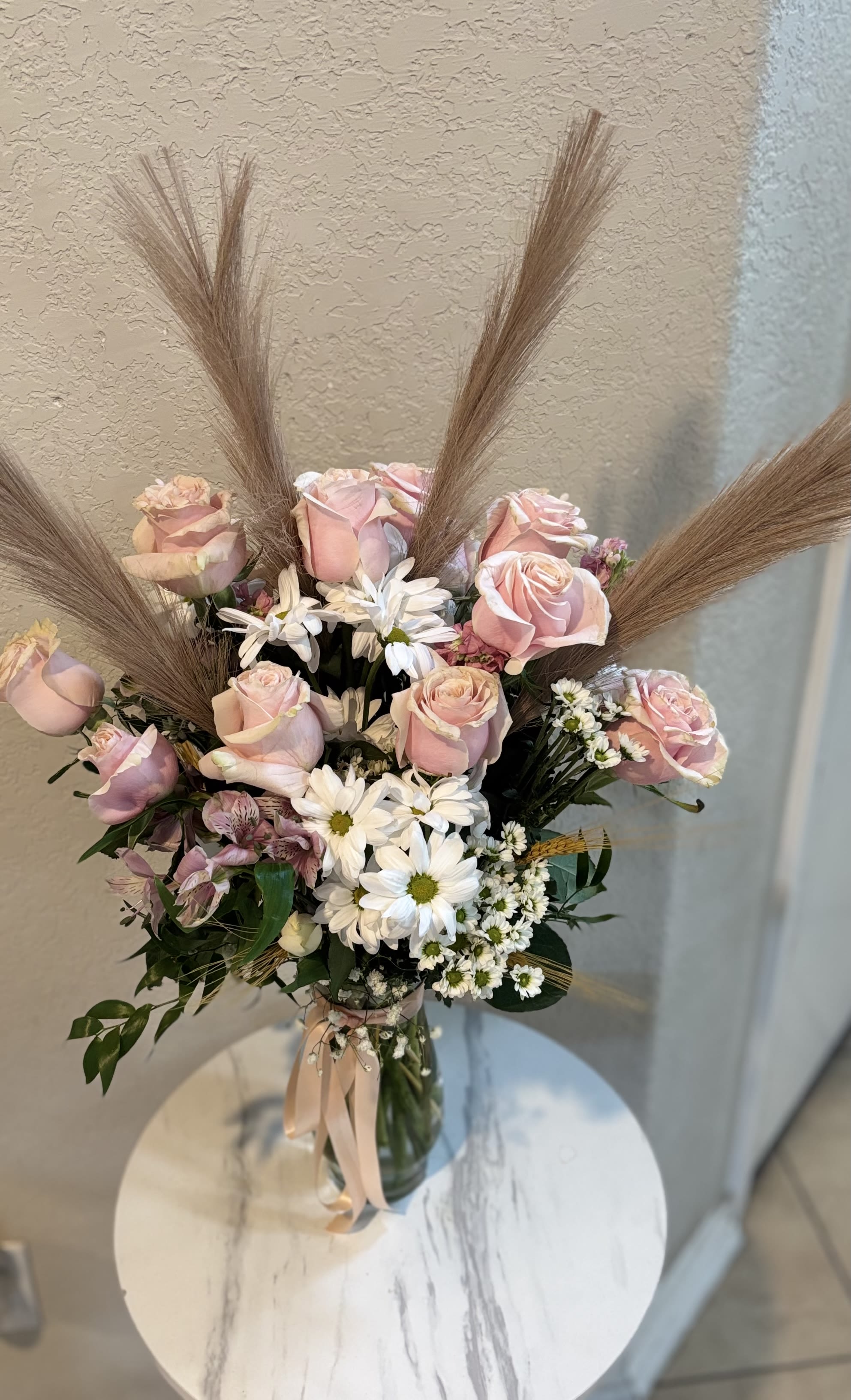 Daisy Boho-Flower Delivery San Antonio - Elegant boho-style arrangement with 12 pink roses, white daisies, baby´s breath and pampas grass in a glass vase, designed by Snyder Flowers, a trusted florist in San Antonio TX. Perfect for birthdays, baby showers or romantic gifts. Same day flower delivery available in San Antonio.