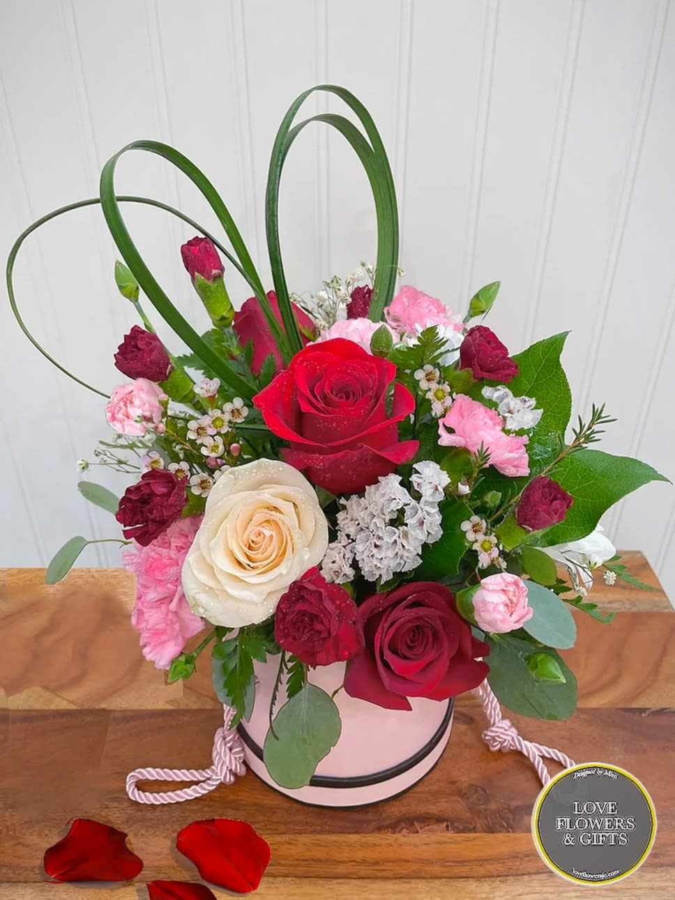 Pink hatbox arrangement with red and cream roses, pink carnations, and decorative grass loops on a wooden surface.
