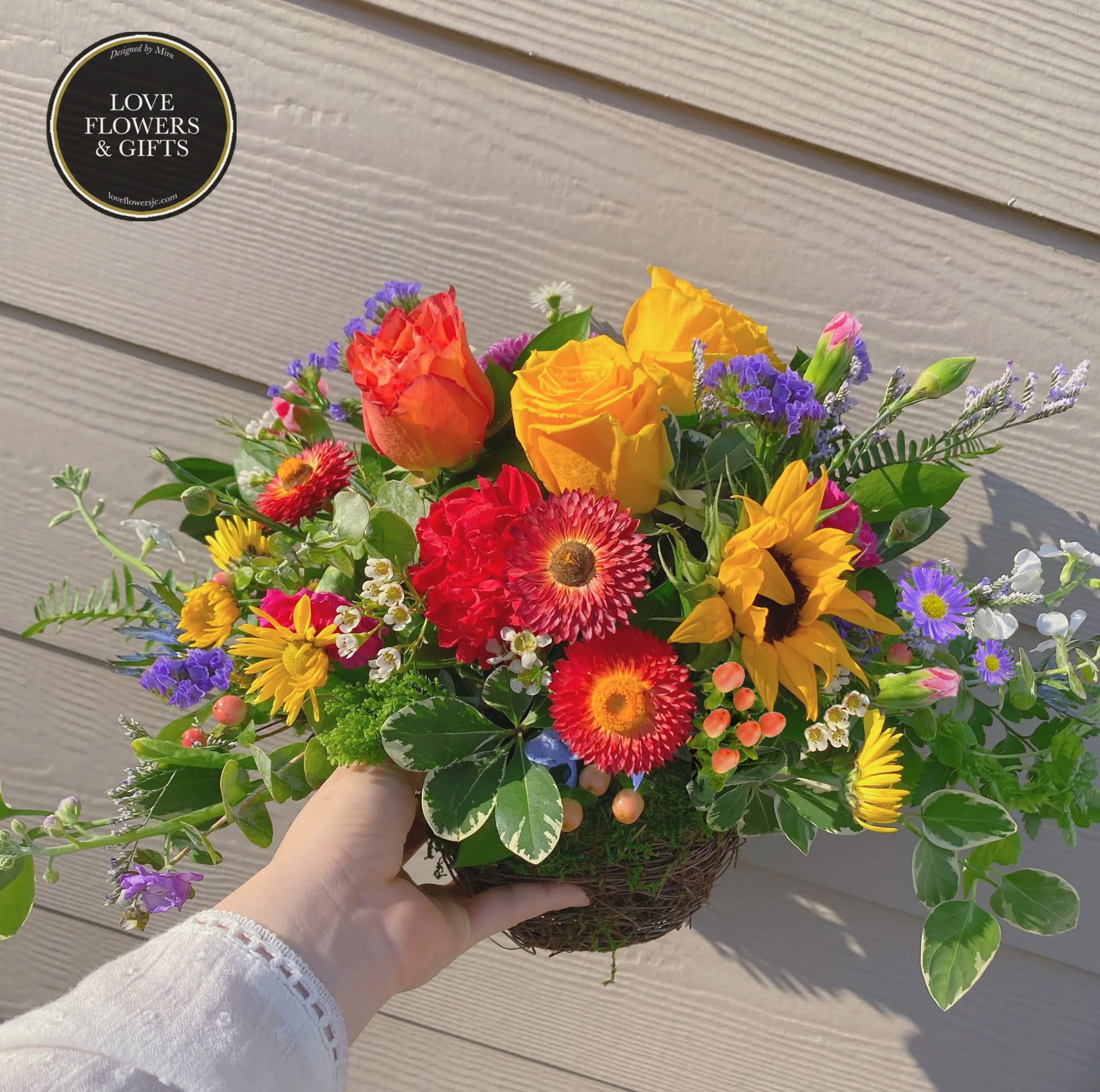 Bright mixed arrangement with yellow roses, red gerberas, and a sunflower in a low twig basket.