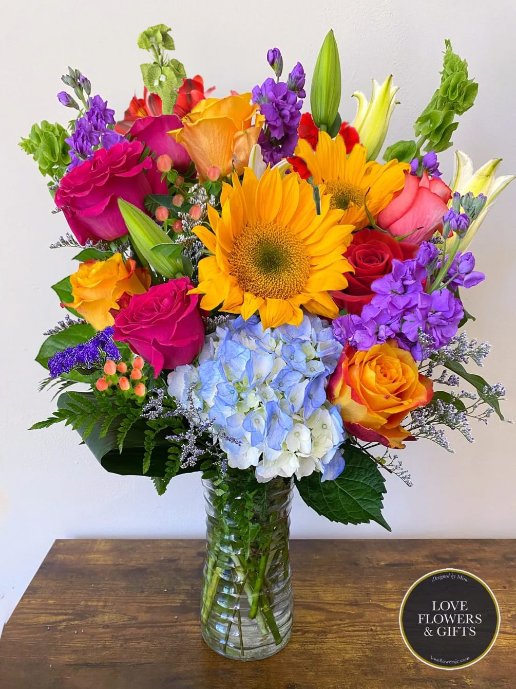 Tall mixed bouquet with sunflowers, roses, lilies, hydrangea, and purple stock in a clear glass vase.
