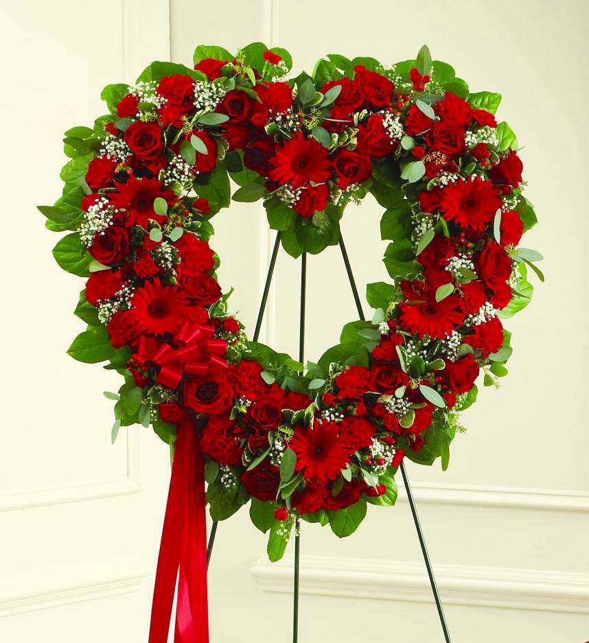 Heart-shaped standing wreath of red roses and other red flowers with a red ribbon.