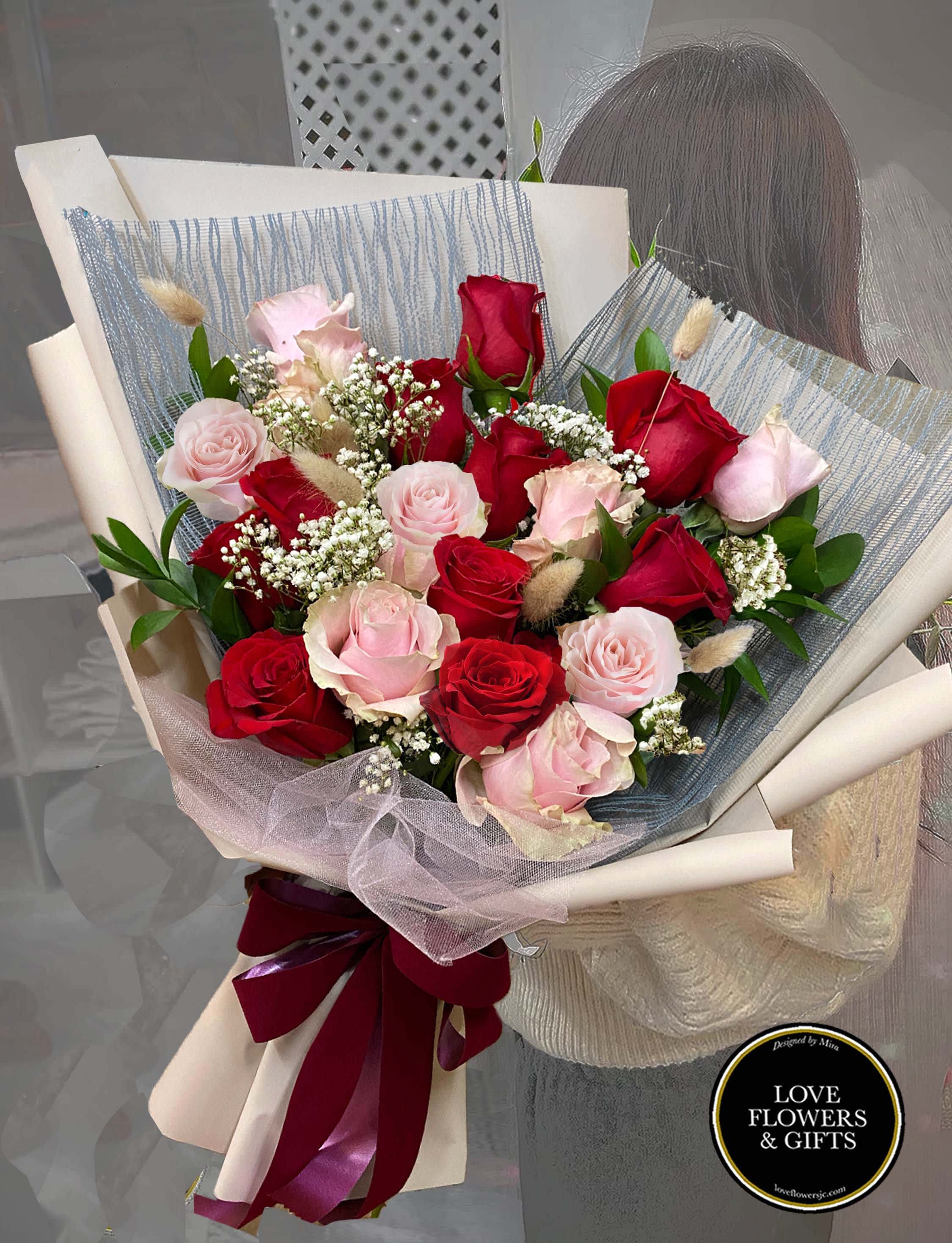 Hand-tied bouquet of red and pink roses with white filler flowers wrapped in cream and gray paper.