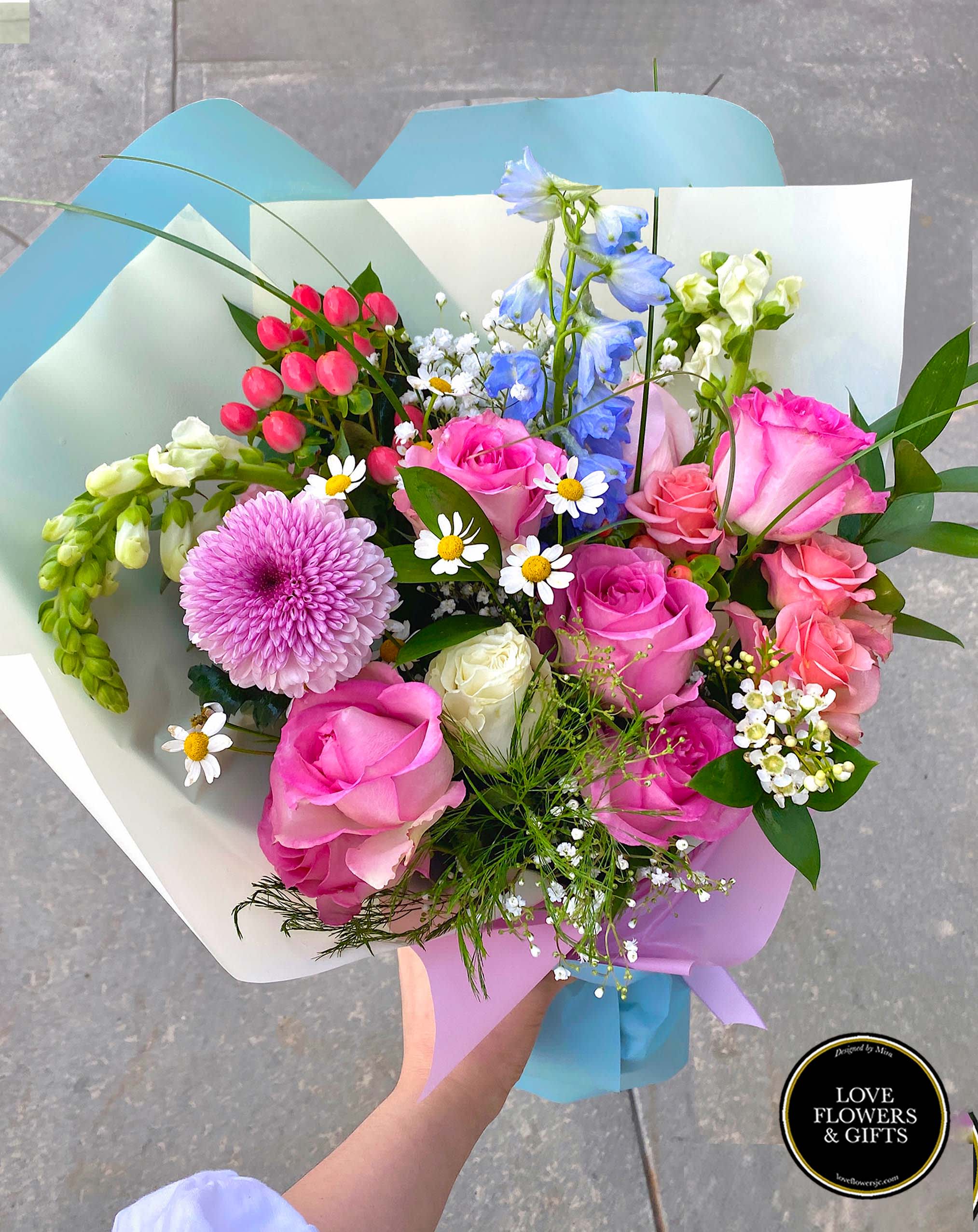 Hand-tied bouquet of pink roses, blue delphinium, white snapdragons, and mixed blooms wrapped in pastel paper