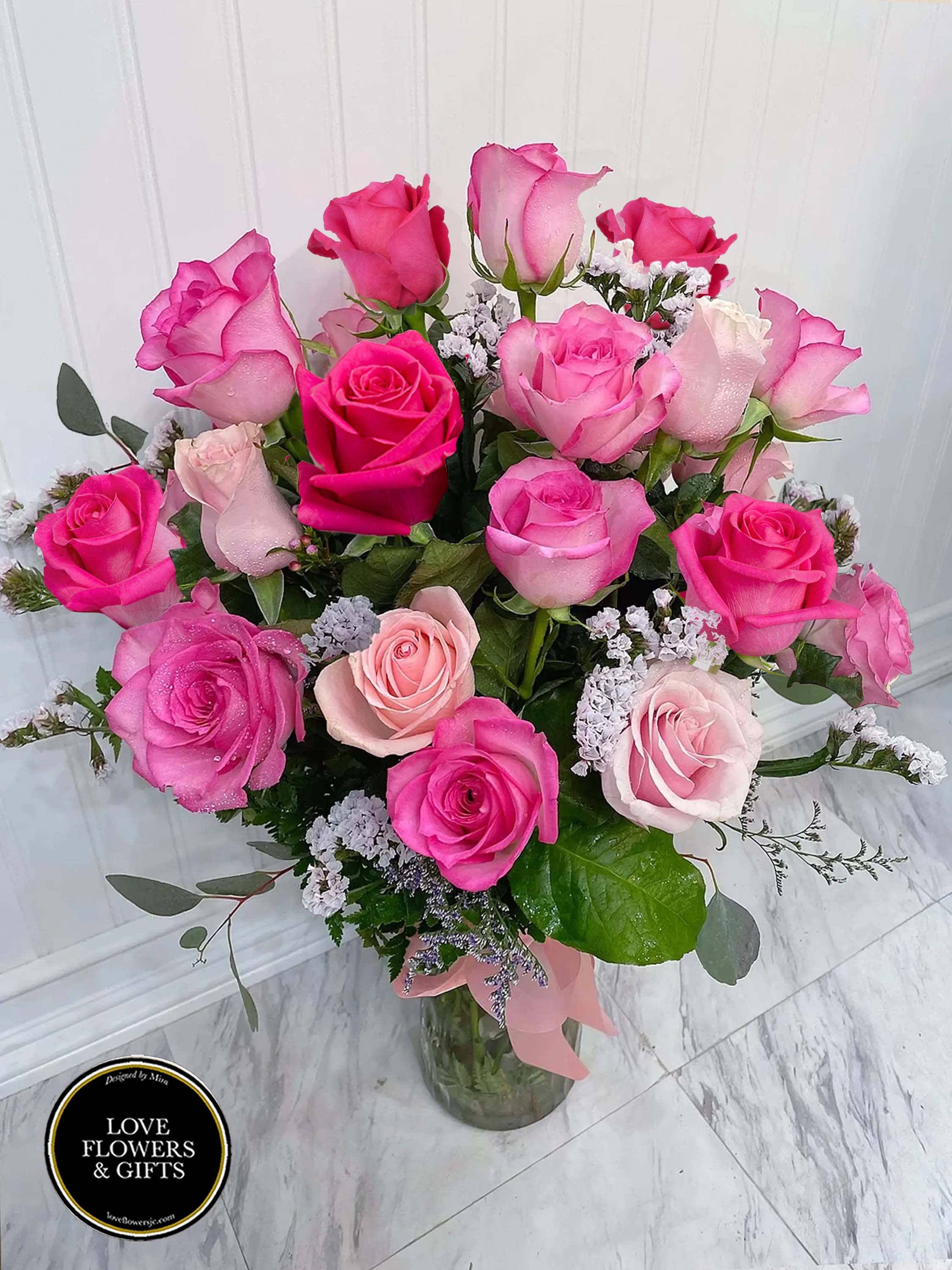 Tall glass vase of hot pink and light pink roses with white filler flowers and a pink ribbon