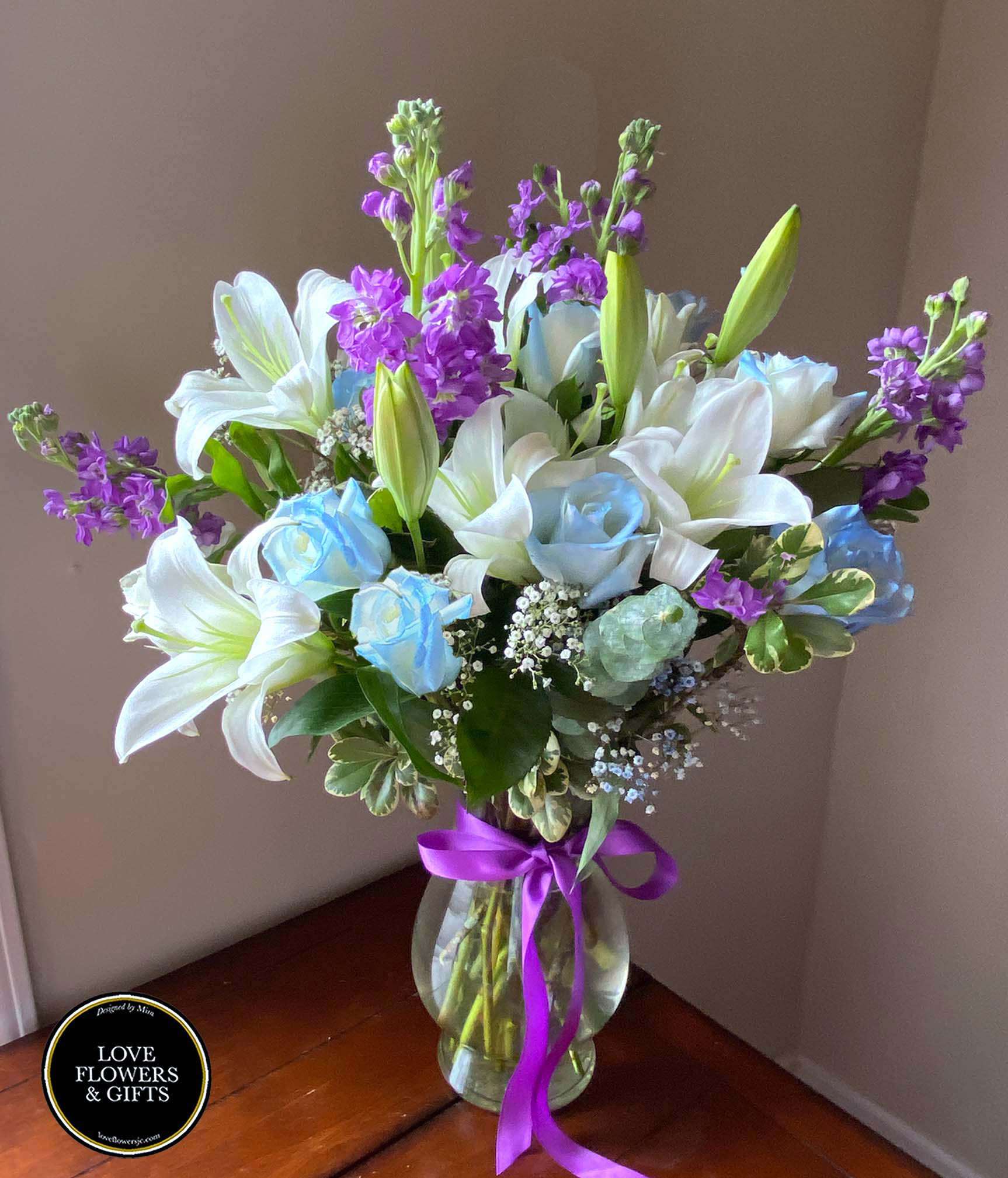 Tall vase arrangement with white lilies, blue roses, and purple flowers tied with a purple ribbon