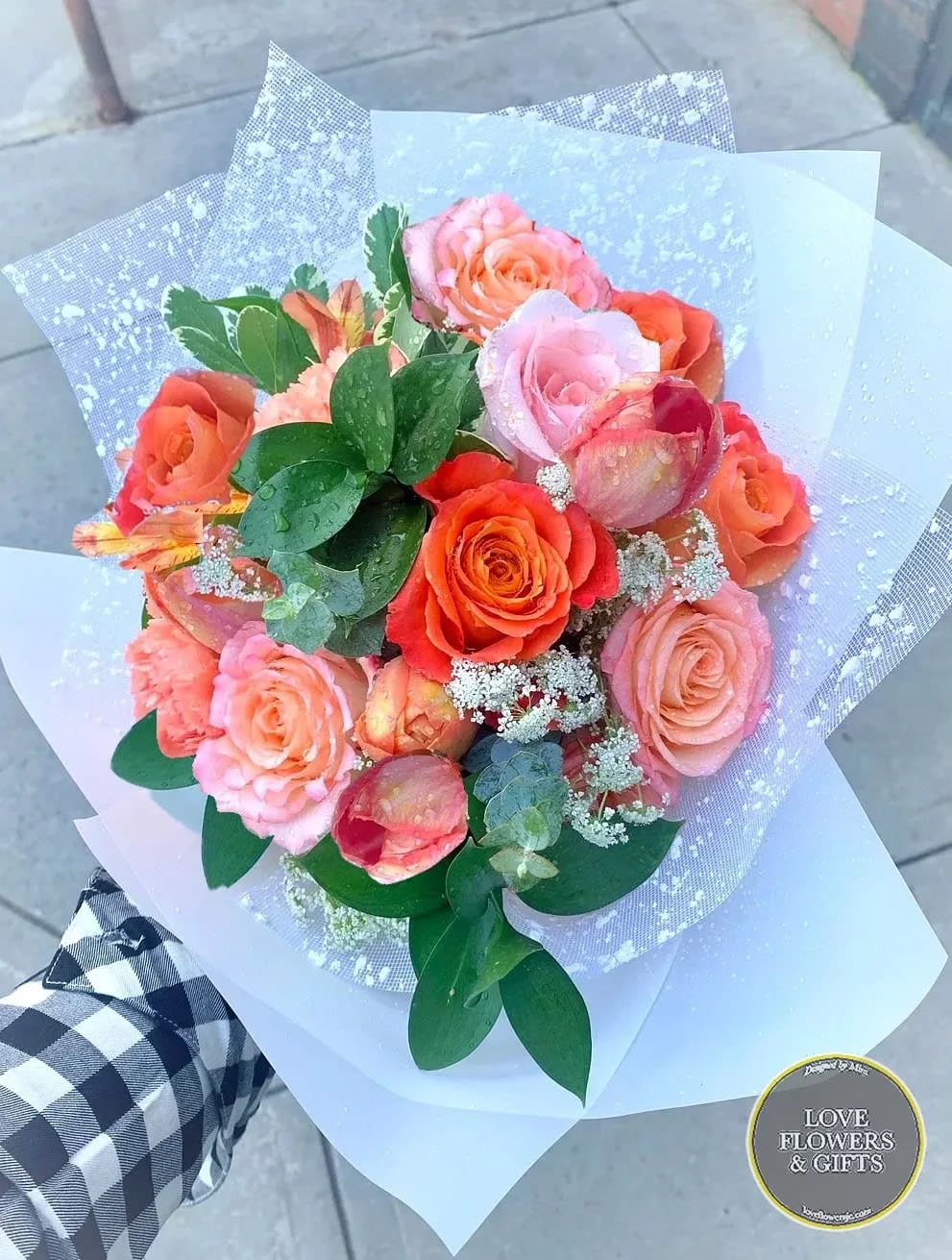 Handheld bouquet of orange and pink roses wrapped in white translucent paper.