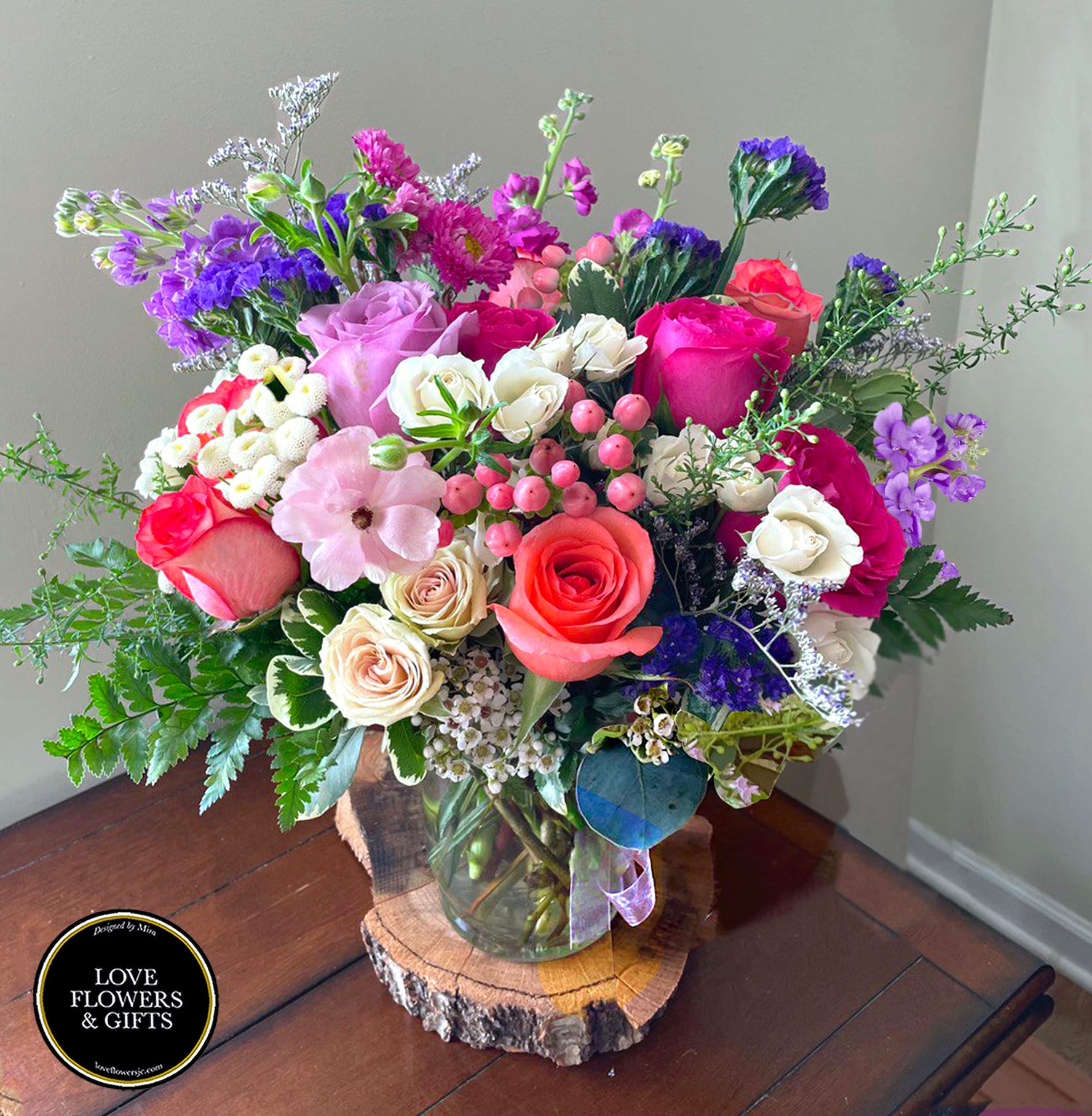 Colorful mixed bouquet of roses and assorted blooms in a clear glass vase on a wood slice.