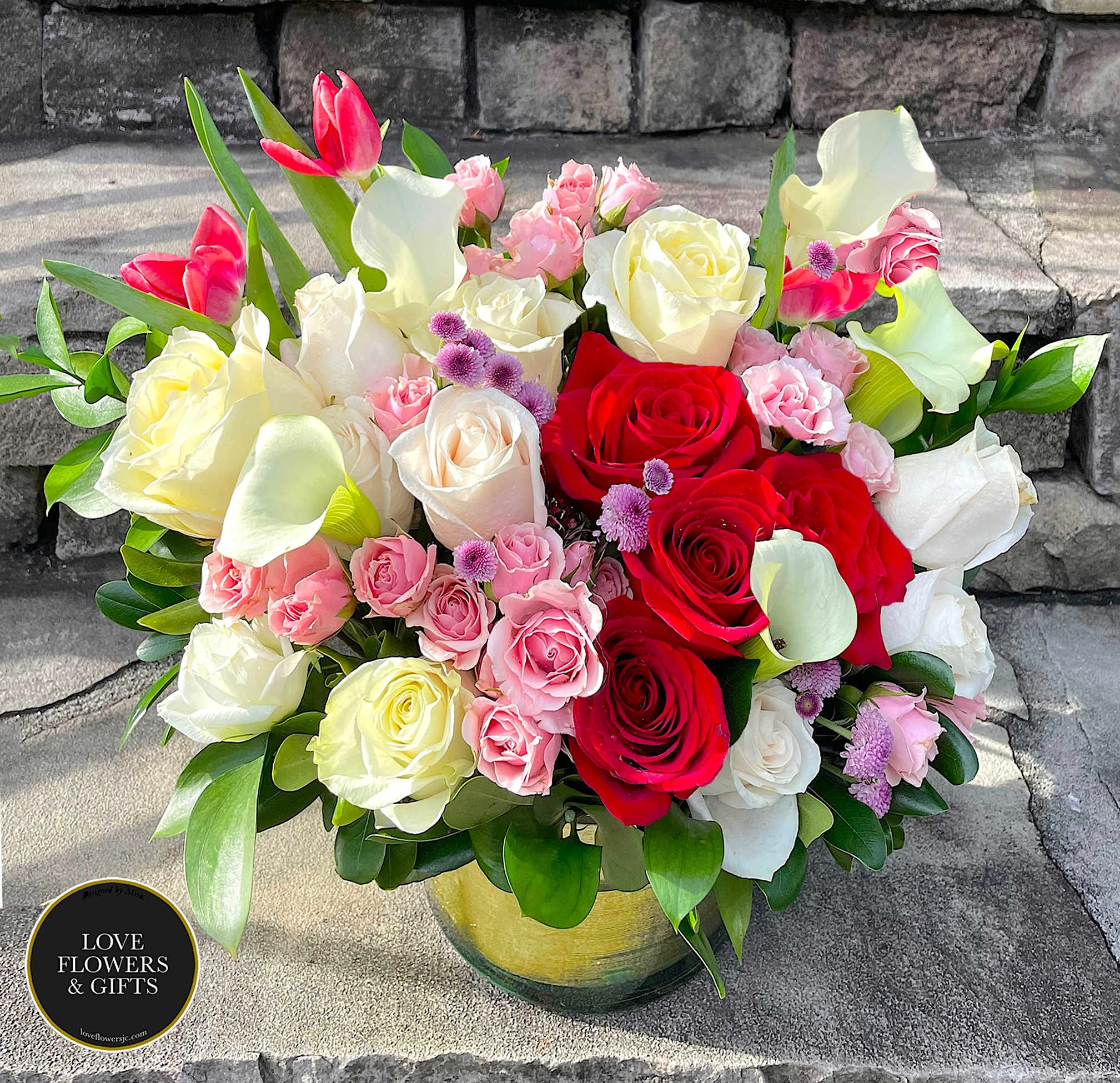 Round arrangement of red and white roses, pink spray roses, and white calla lilies in a glass vase.