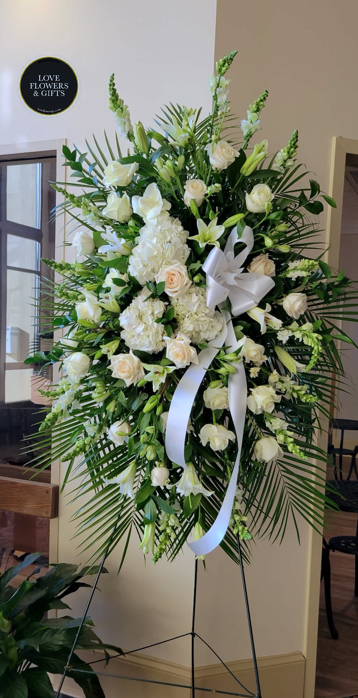 Tall standing spray of white roses, lilies, hydrangeas, and palm fronds with a white bow on an easel.