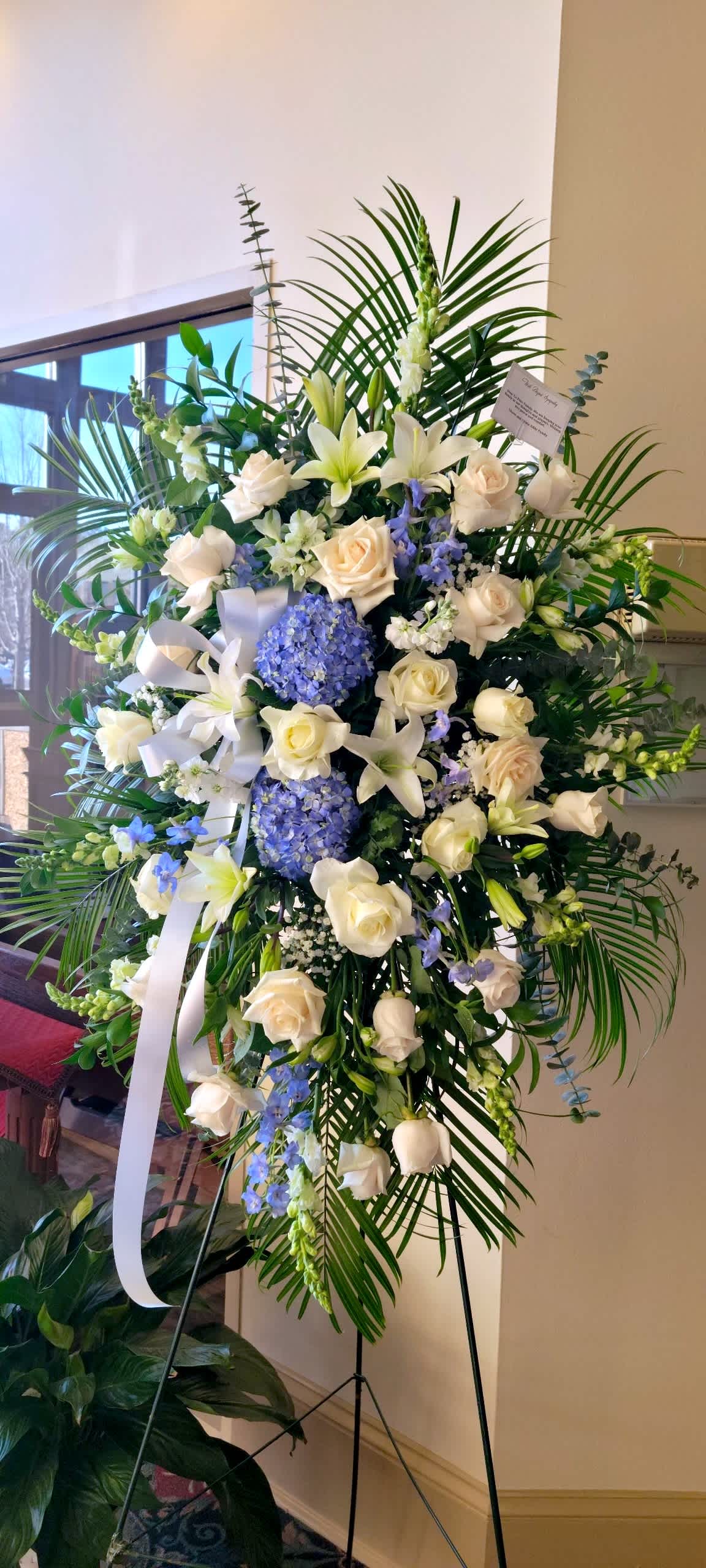 Tall standing spray of white roses, lilies, and blue hydrangeas with palm fronds on an easel.
