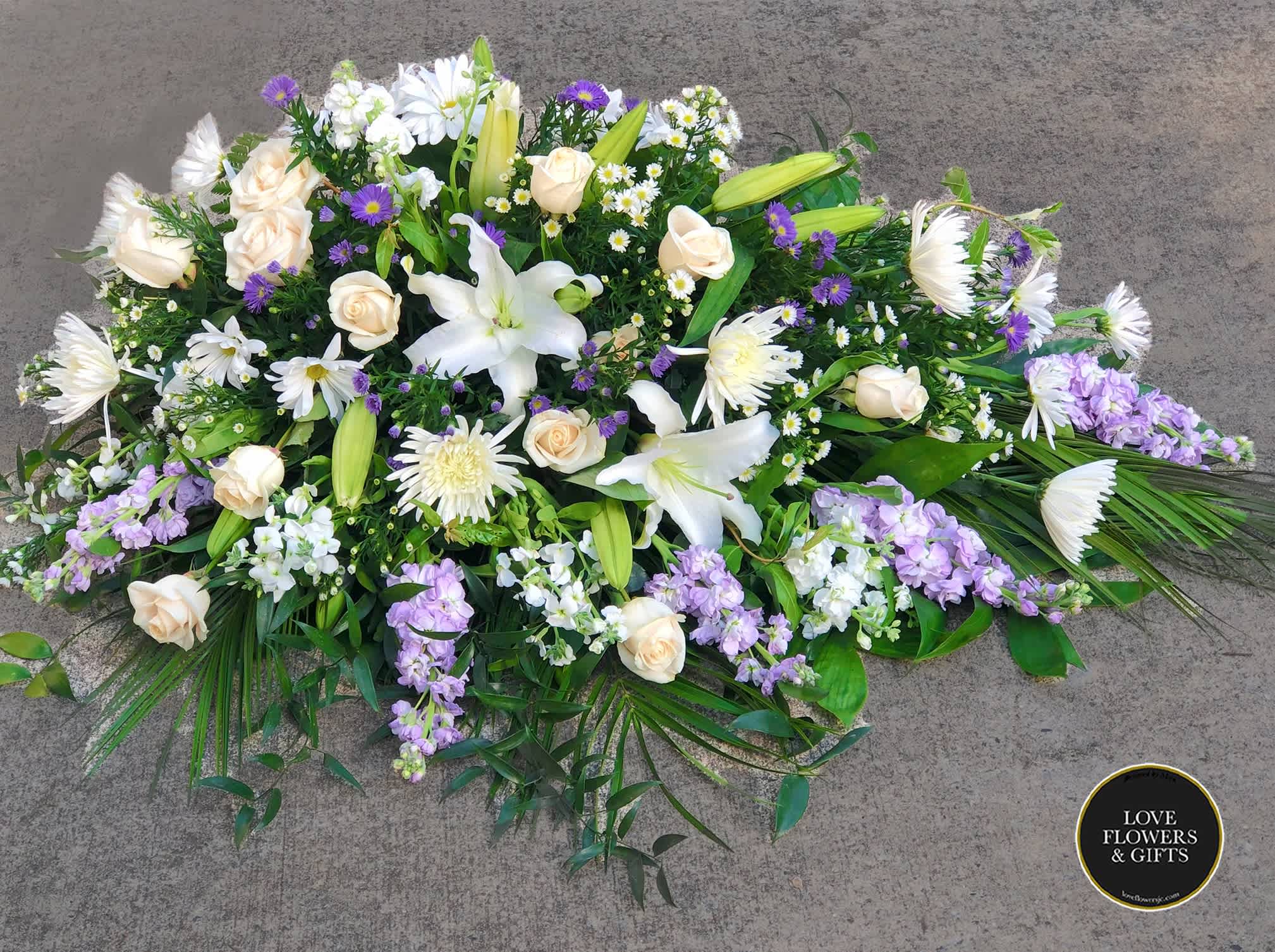 Purple and White (Half Casket Spray)  - This large half casket spray is made to remember her, featuring a variety of white and lavender flowers and greenery. This half casket is for a partially open casket. (Please add the funeral service time at checkout, we deliver before then)  If you like us to change or add certain colors, please write in "Florists Instructions" at Checkout.