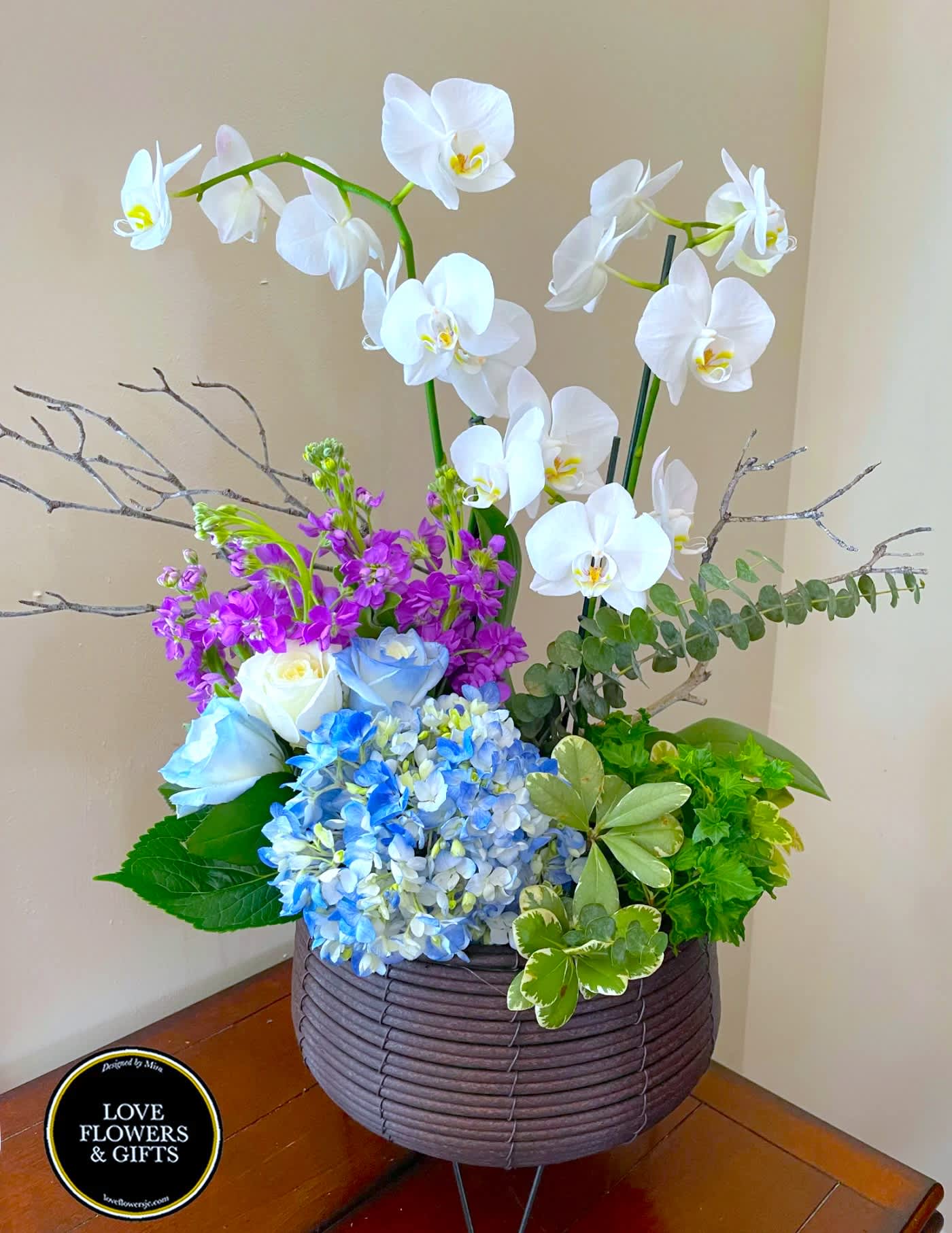 Tall arrangement of white orchids, purple flowers, blue hydrangeas and white roses in a brown basket container.