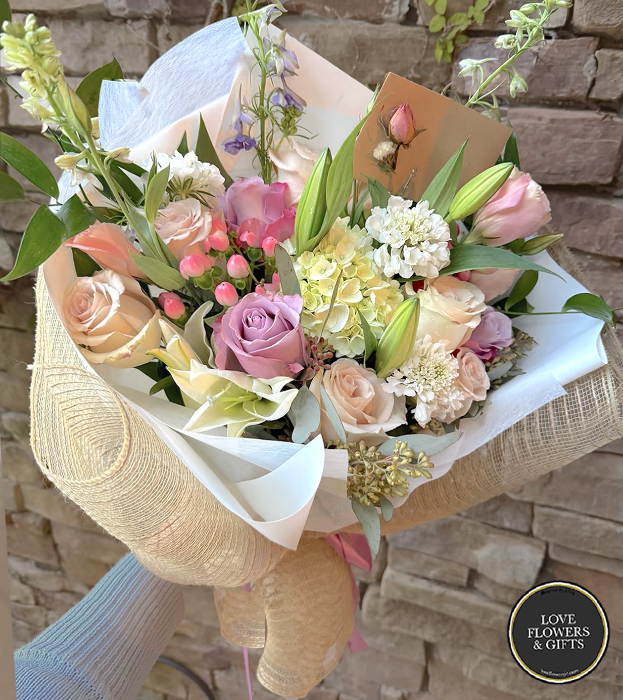 Hand-tied bouquet of pastel roses, lilies, and mixed blooms wrapped in white and burlap-style paper