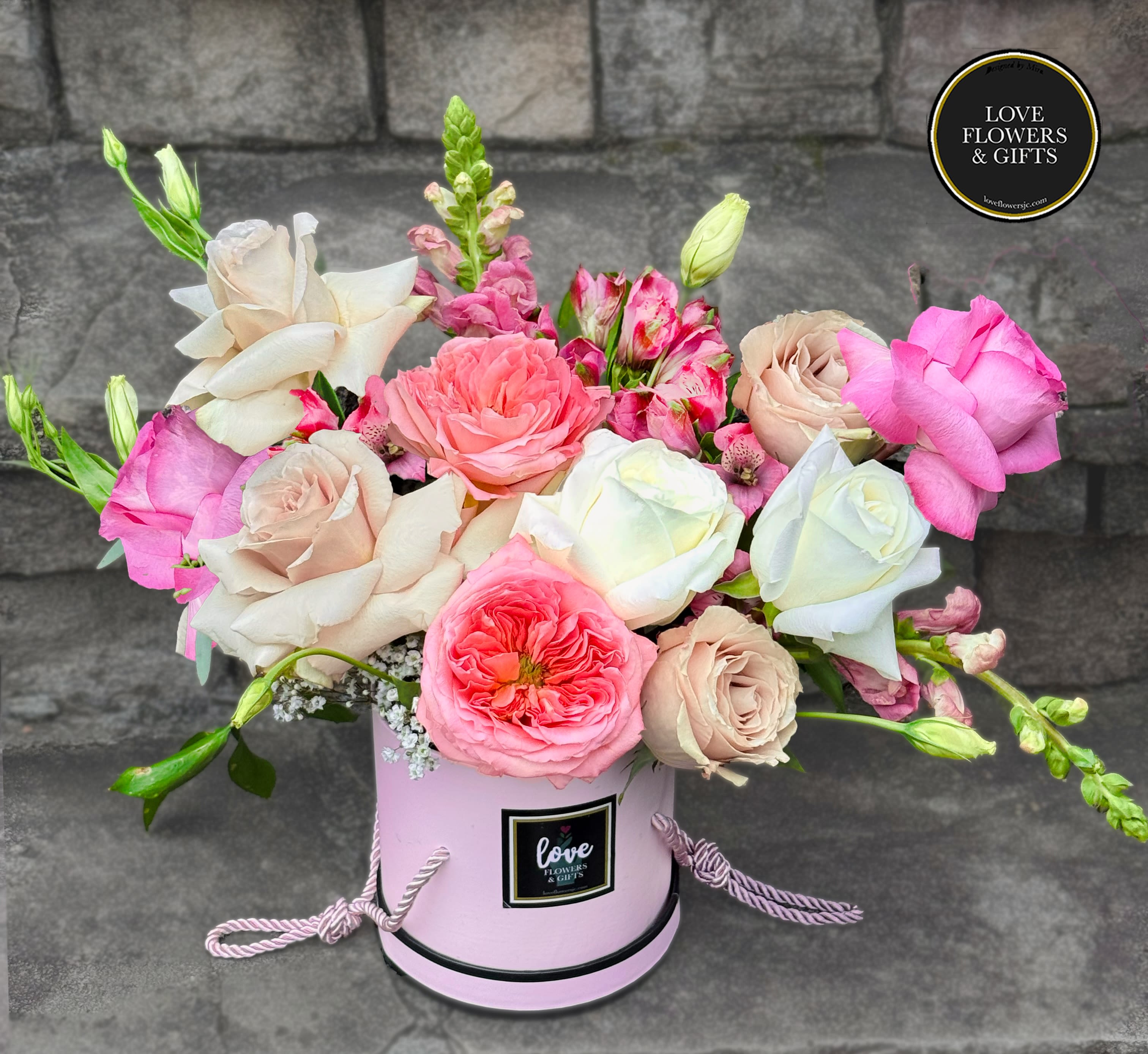 Pink and cream roses with mixed blooms arranged in a round pink hatbox-style container.