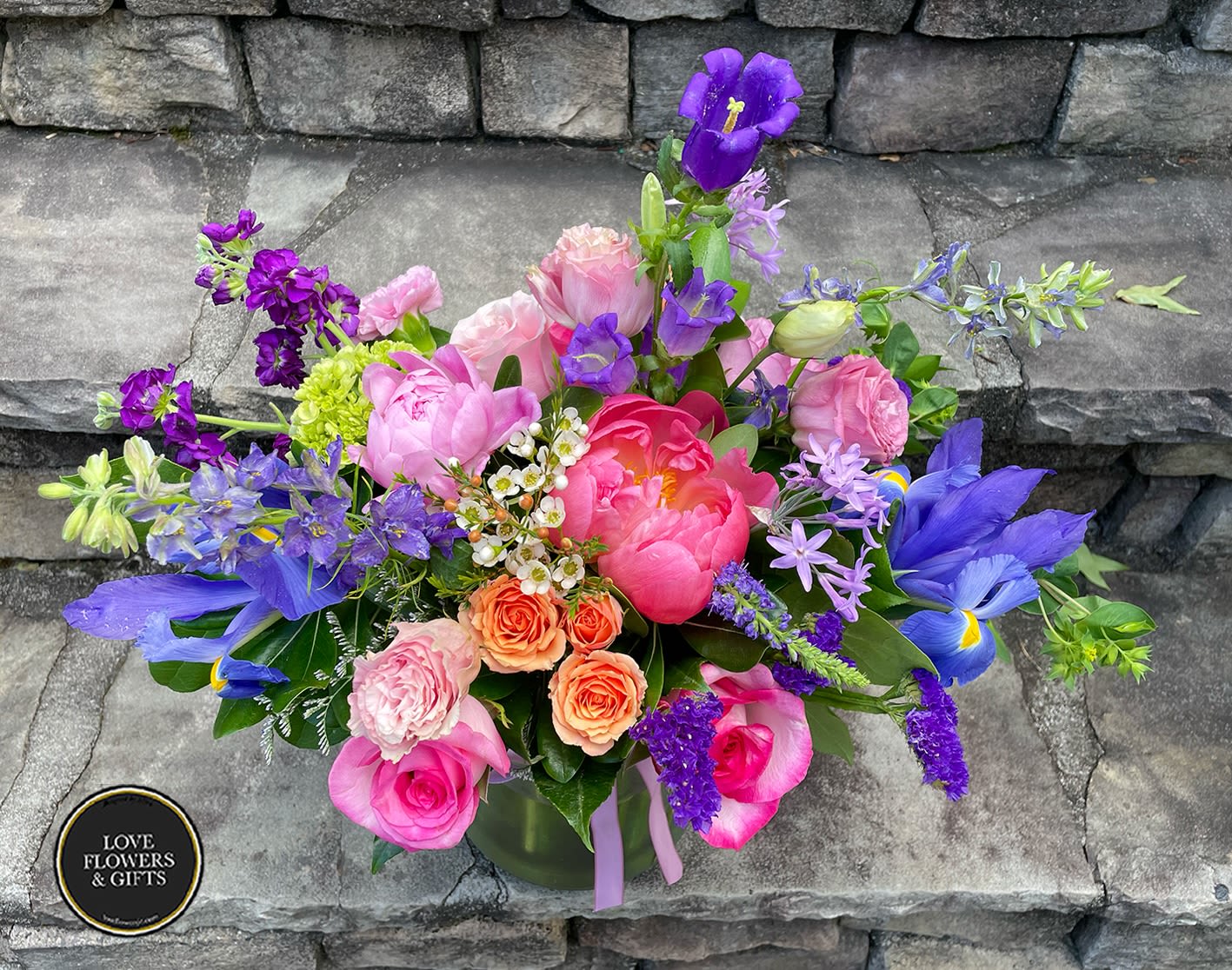 Vibrant mixed arrangement of pink peonies, roses, and purple irises in a clear glass vase