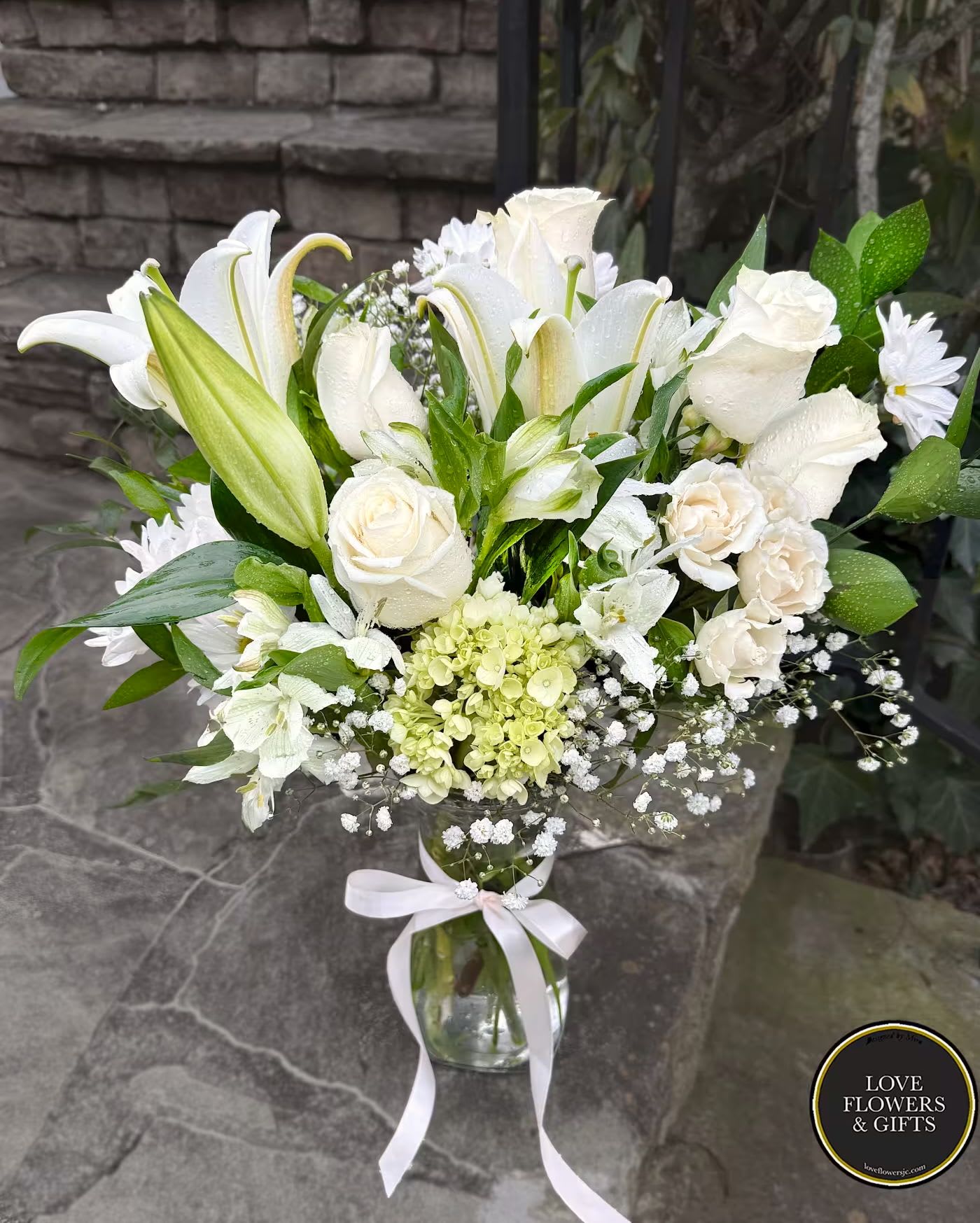 Tall white flower arrangement in a clear glass vase with a white ribbon bow