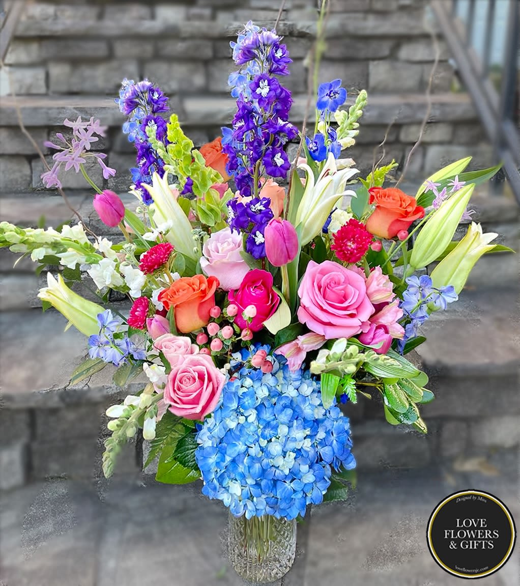 Tall mixed bouquet with pink roses, lilies, blue hydrangea, and purple delphinium in a clear vase