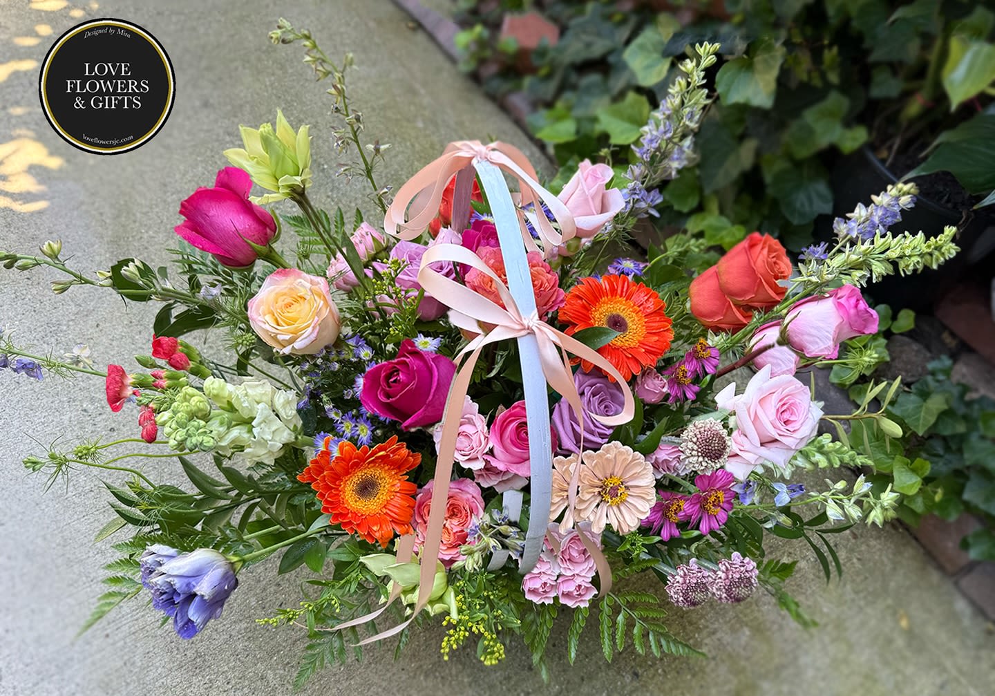 Colorful basket arrangement of pink roses, orange gerbera daisies, and mixed blooms with pastel ribbon bows