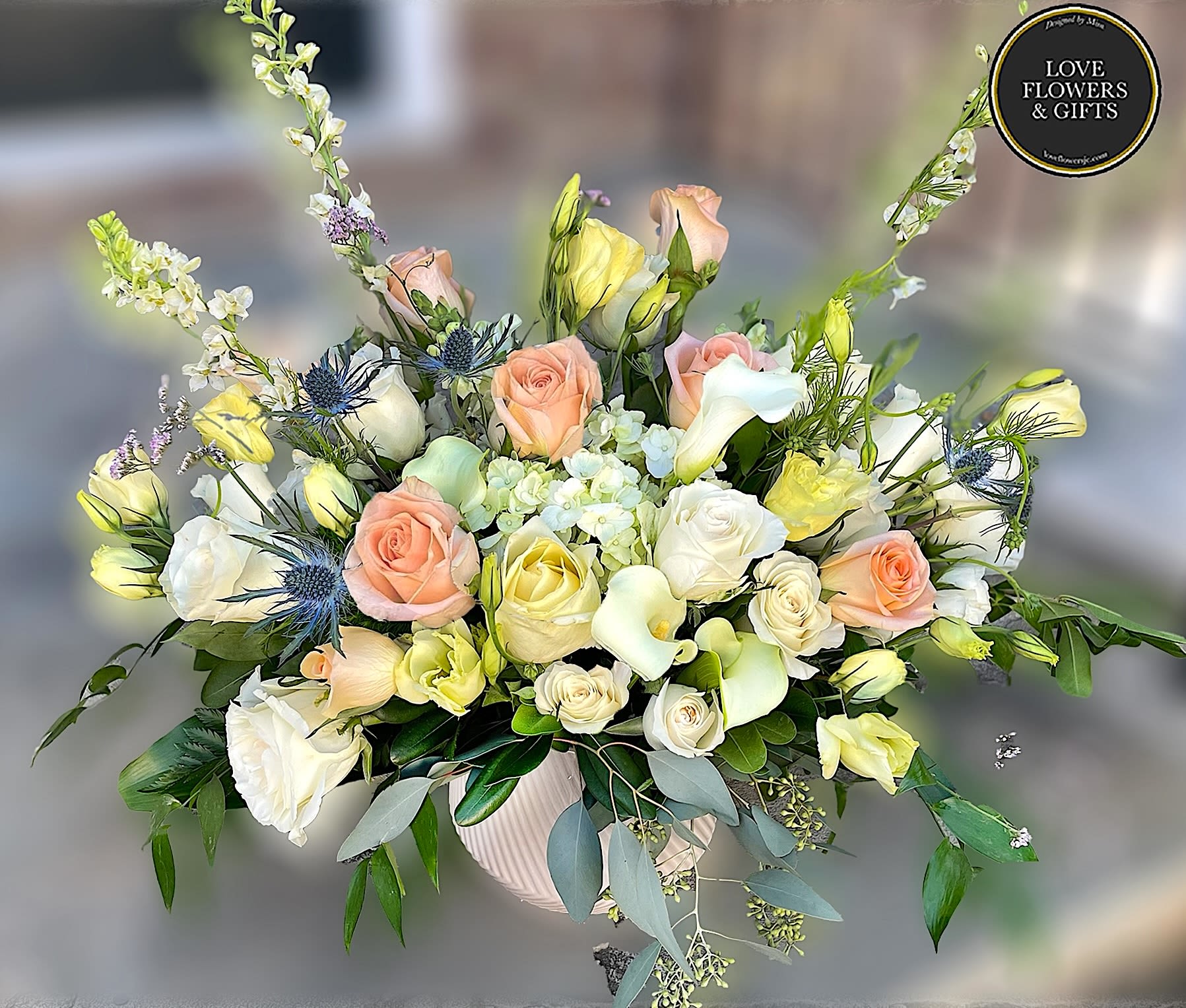 Low ceramic bowl of white, peach, and yellow roses, calla lilies, and mixed blooms in a lush arrangement.