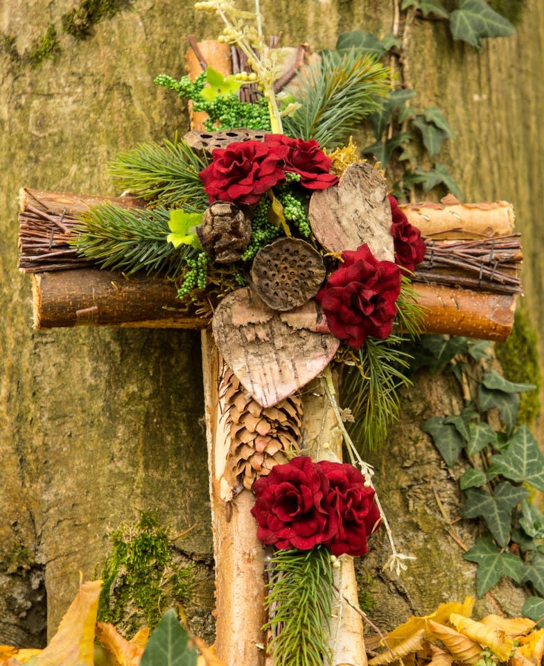 Rustic wooden cross decorated with red flowers, evergreens, bark hearts, and pinecones.